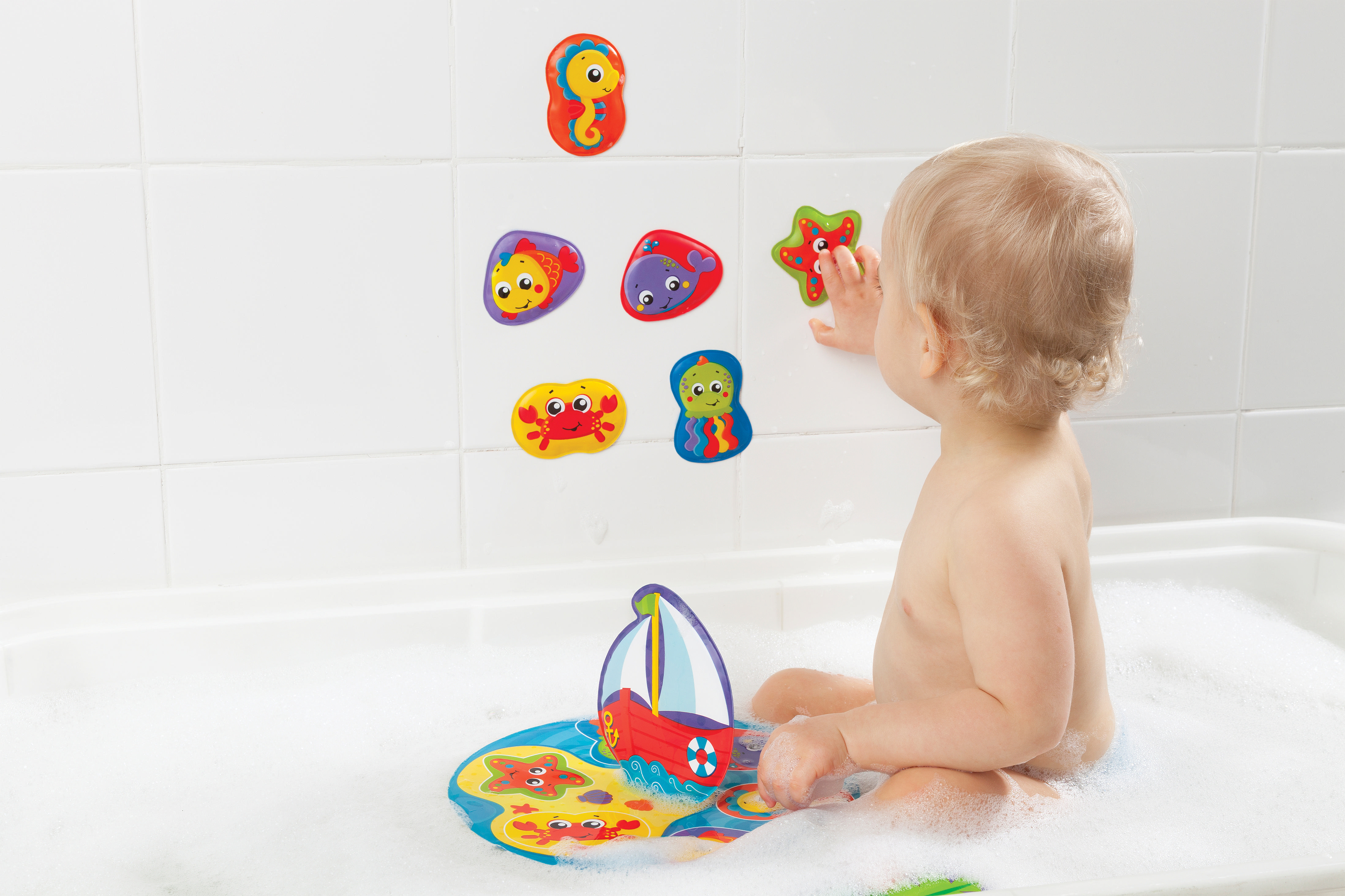 Baby in einer Badewanne mit Meeresbewohner-Spielzeug an der Wand und einer bootsförmigen Matte.