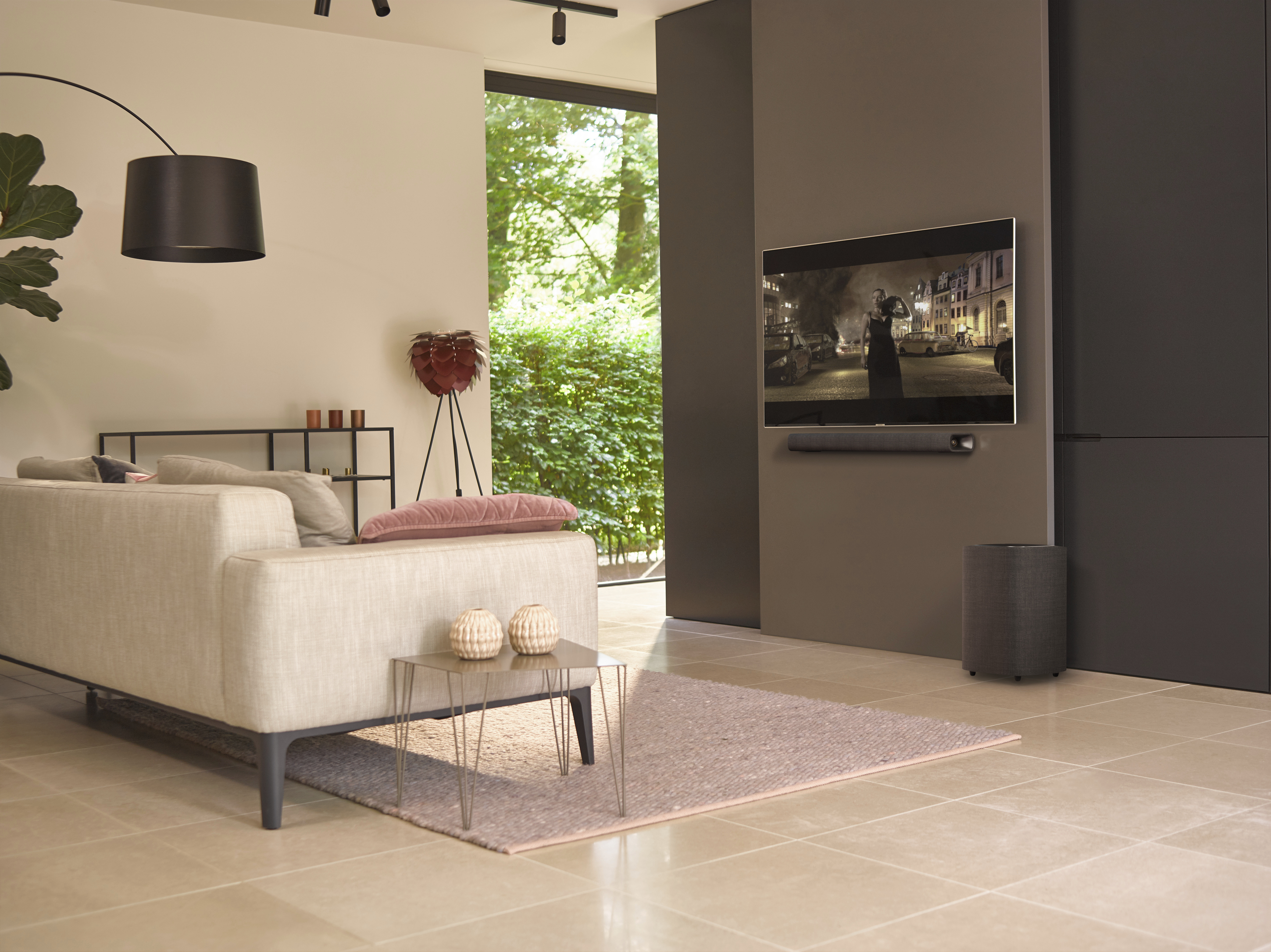Woonkamer met bank, tv aan de muur, soundbar en subwoofer. Groot raam met groen uitzicht.