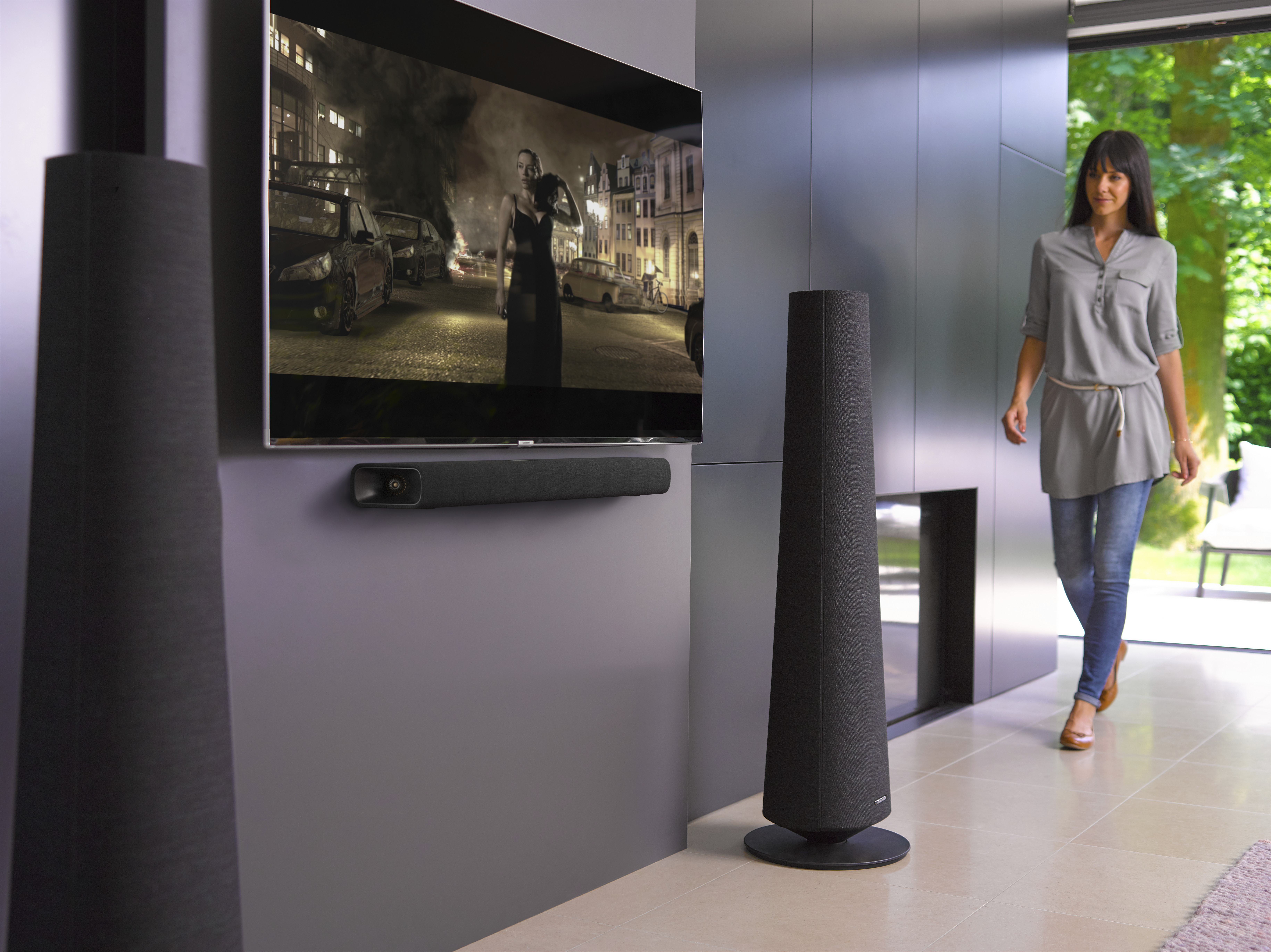 Een vrouw loopt langs een tv met een filmscène, twee luidsprekers en een soundbar.