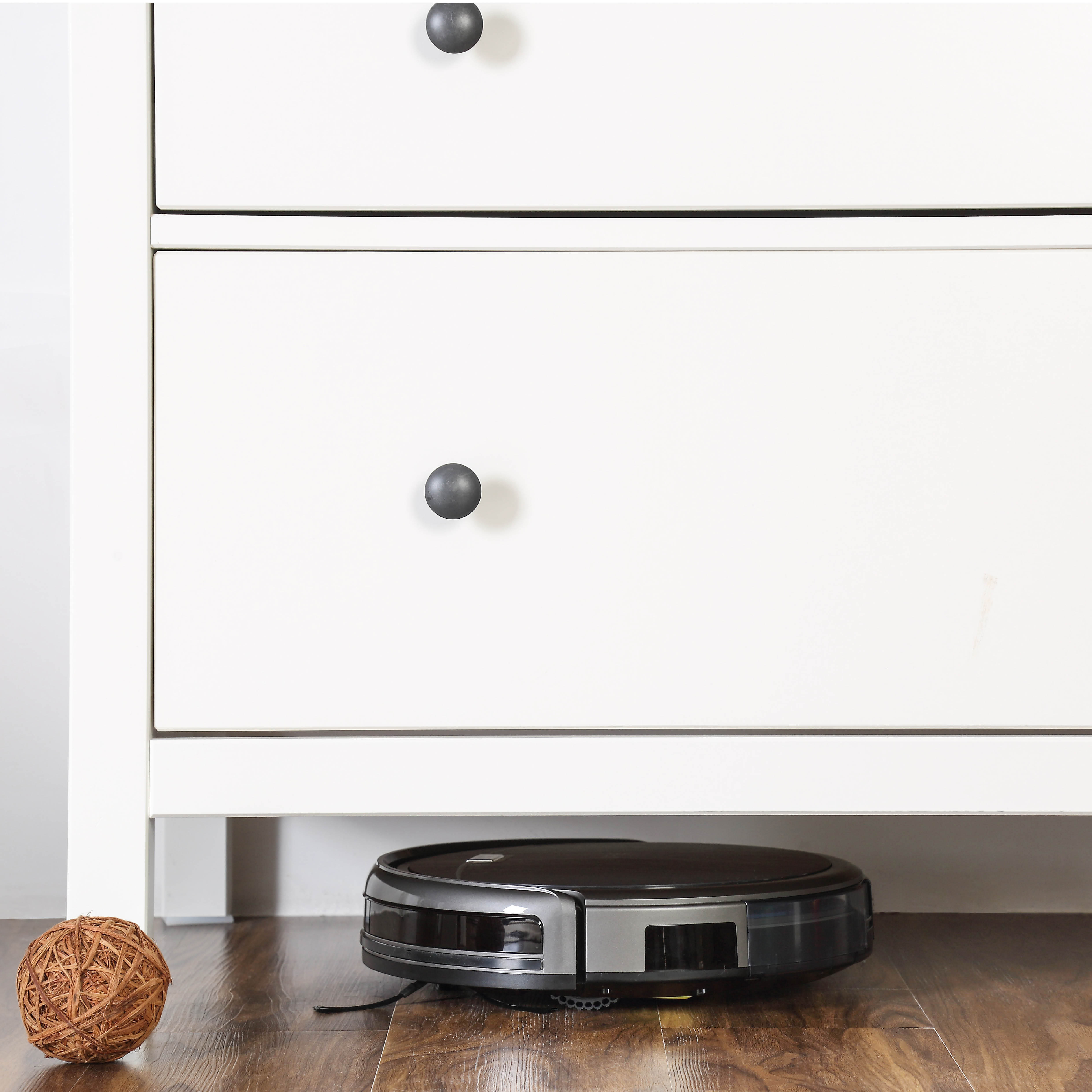Roboter-Staubsauger unter weißem Schrank mit Holzkugel auf dem Boden. Hintergrund ist weiße Wand.