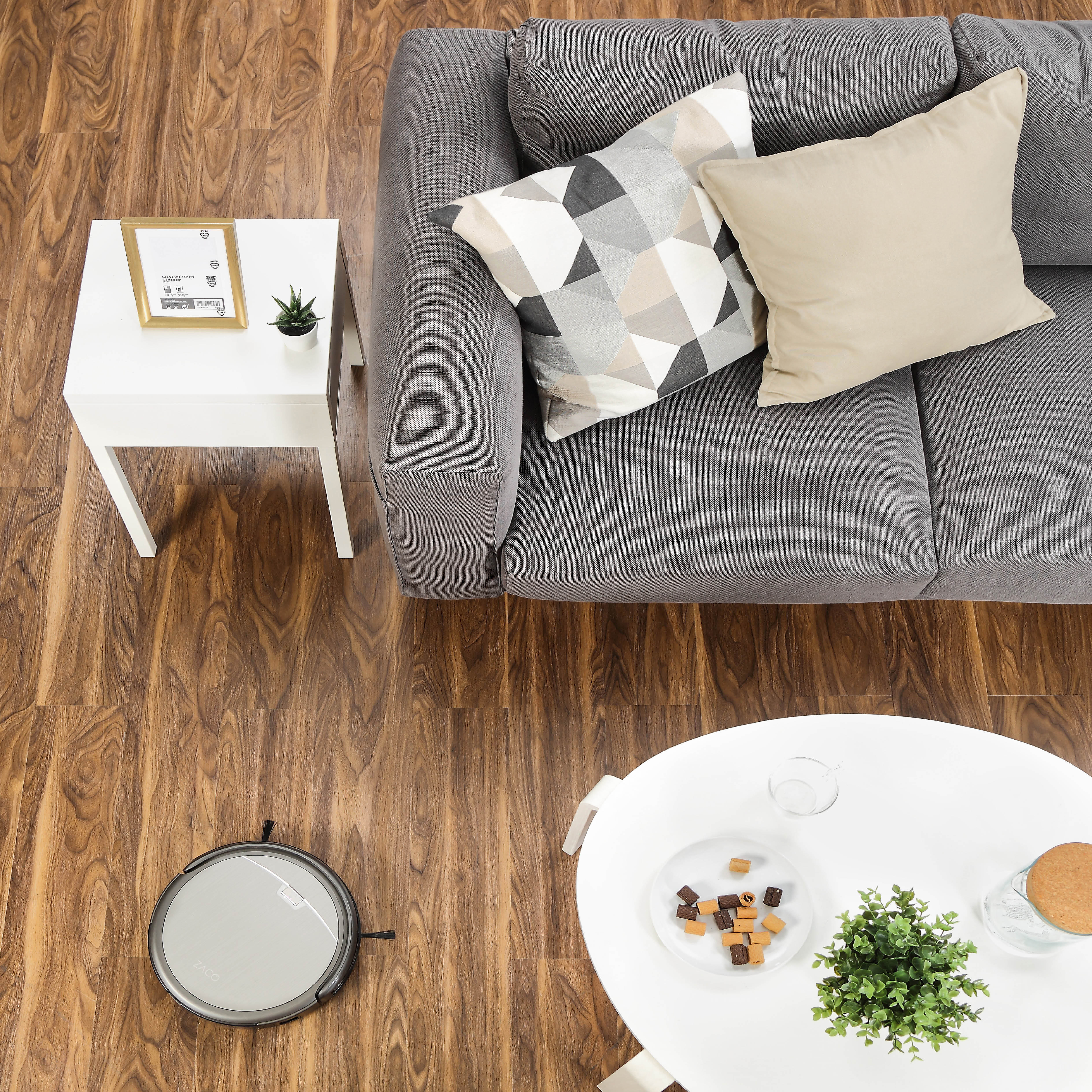 Draufsicht auf einen Raum mit grauem Sofa, weißen Tischen und einem Roboter-Staubsauger.