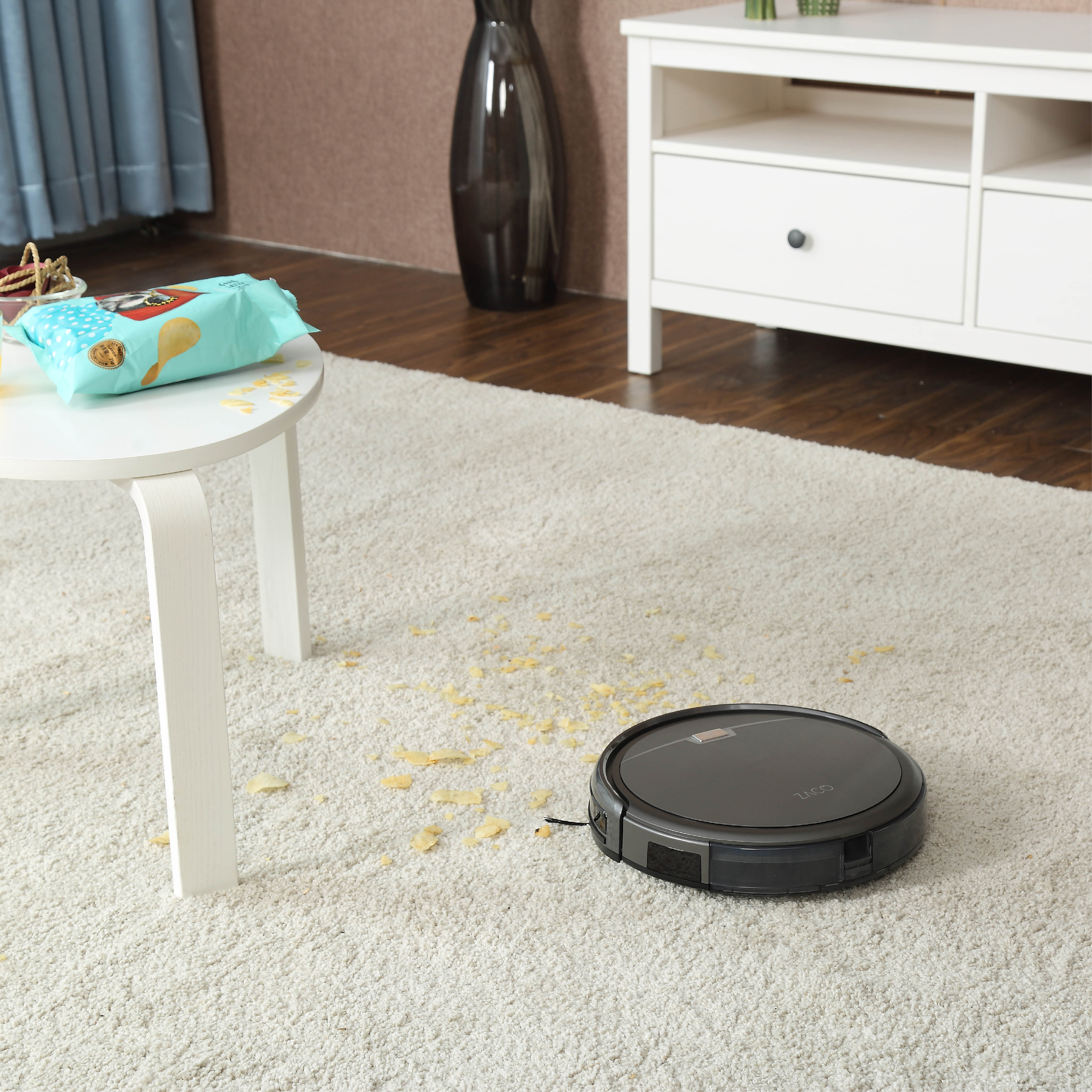 Roboter-Staubsauger auf Teppich. Chips auf dem Boden, Tisch mit Snacks und weißer Schrank.