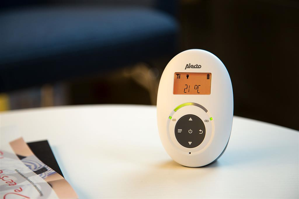 White baby monitor. Oval shape, screen showing temperature. Buttons and a green light. Placed on a white table.
