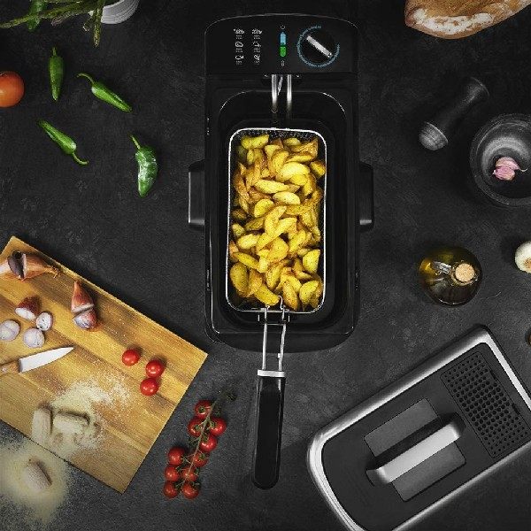 Vista superior de freidora con patatas, verduras y accesorios sobre superficie negra.