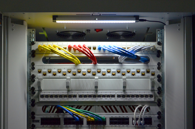 Server-Rack mit Netzwerkkabeln. Gelbe, rote und blaue Kabel sind sichtbar. Weißes Panel mit Anschlüssen.