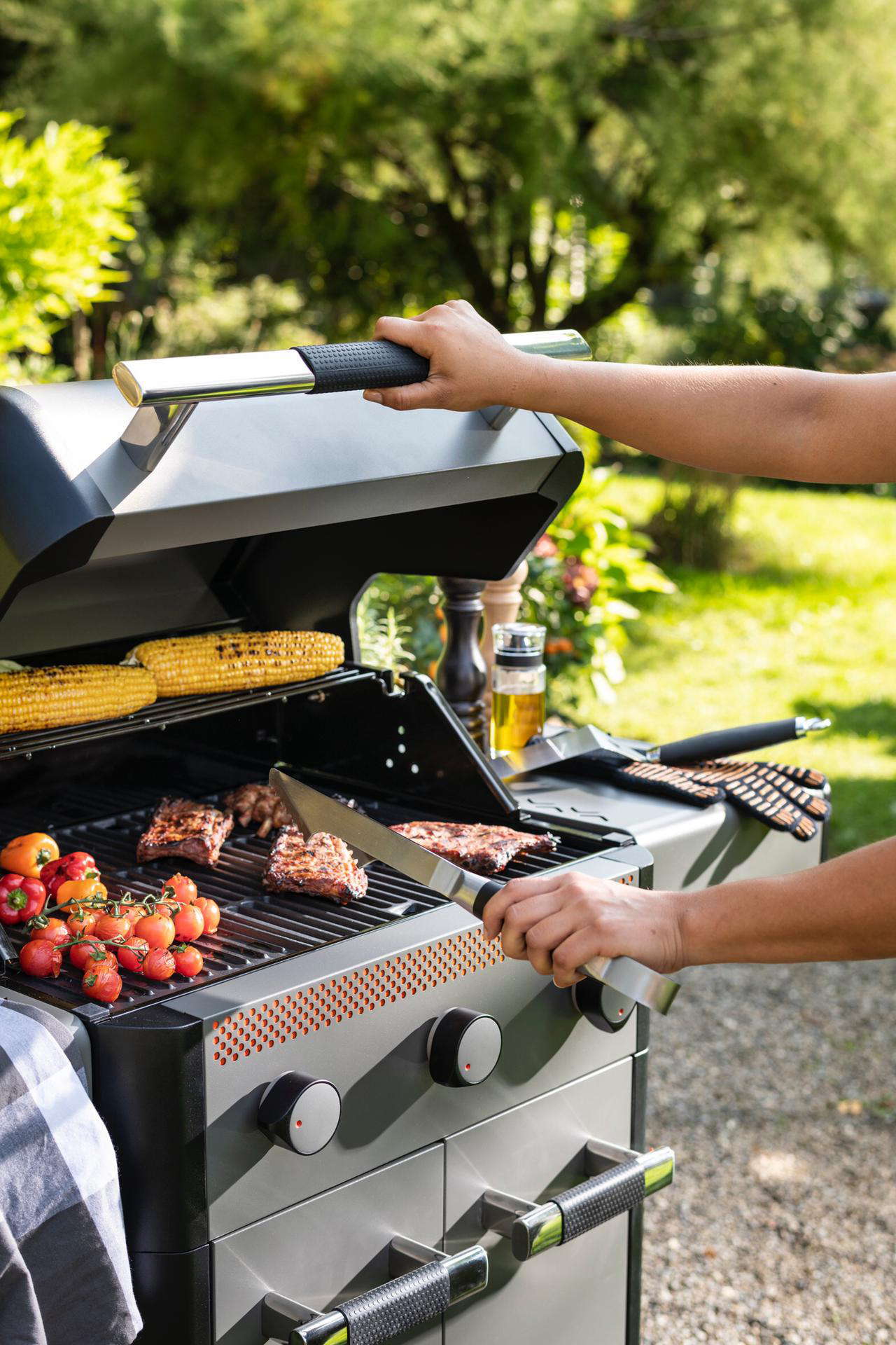 Person grillt Essen auf einem Grill. Mais, Tomaten und Steak. Draußen, sonnig.