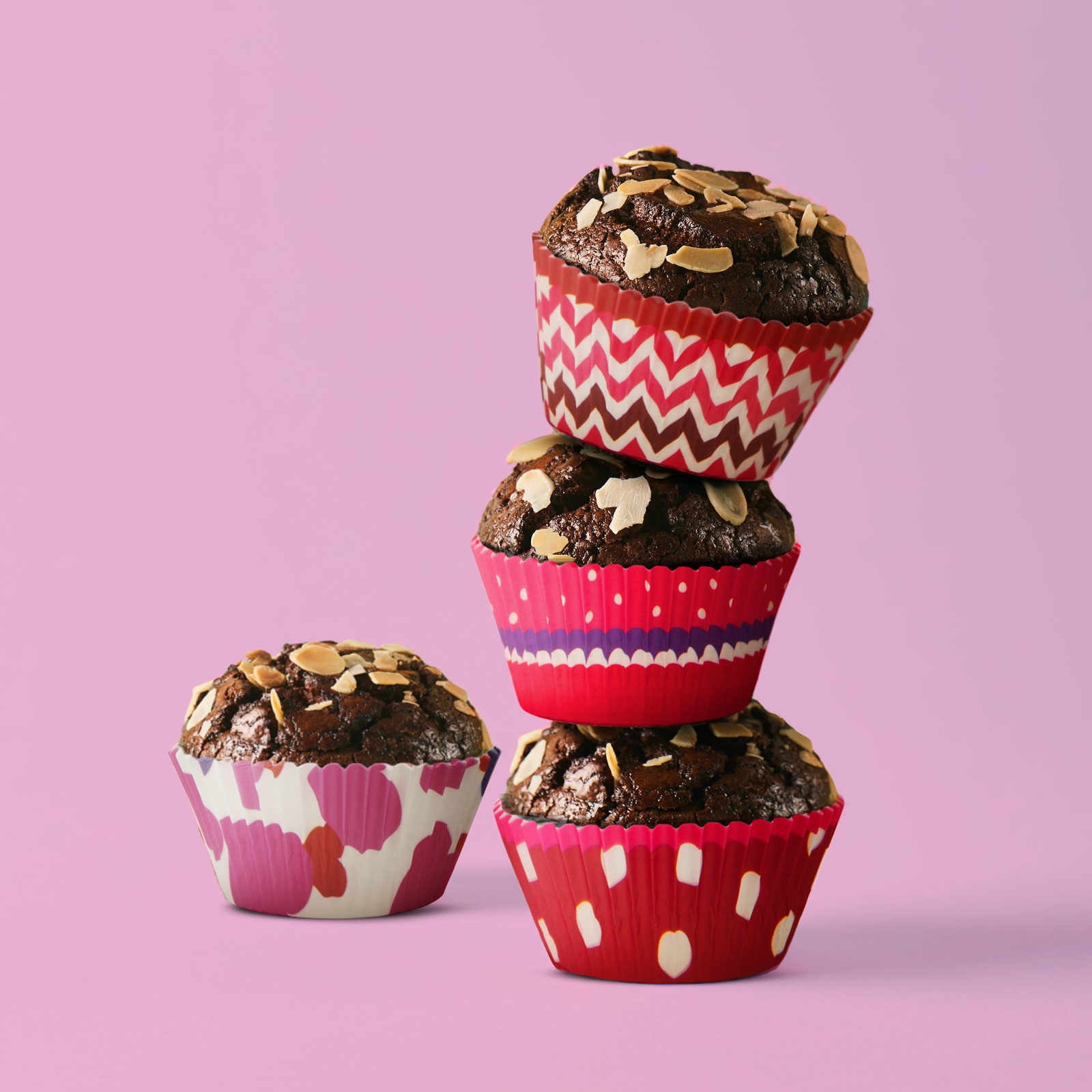 Négy muffin színes papírpohárban, mandulával megszórva, rózsaszín háttérrel.