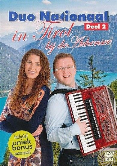 Man speelt accordeon, vrouw ernaast, bergen en meer op achtergrond.