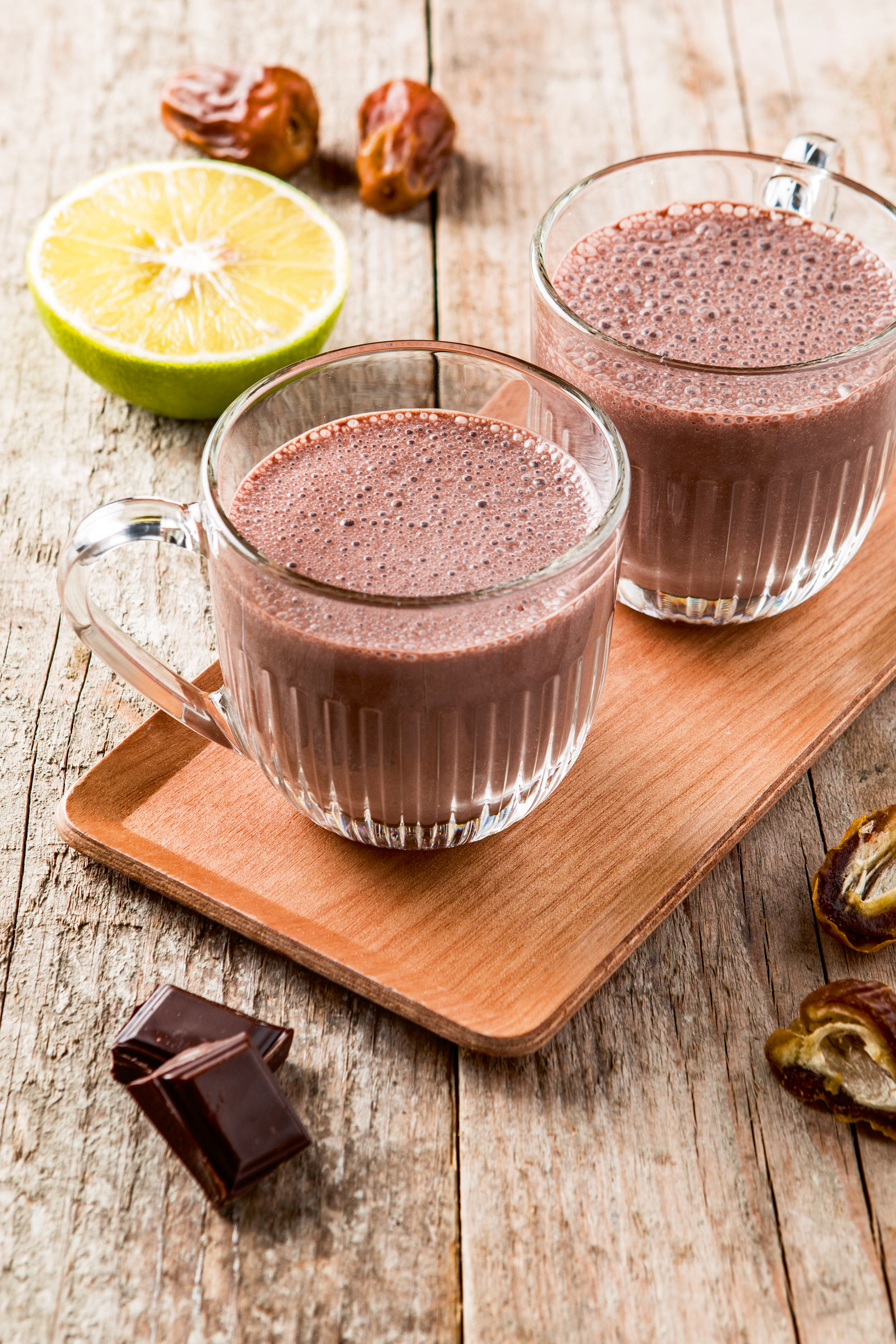 Zwei Schokoladenmilchshakes in Gläsern auf einem Holztablett, mit Limette und Datteln.