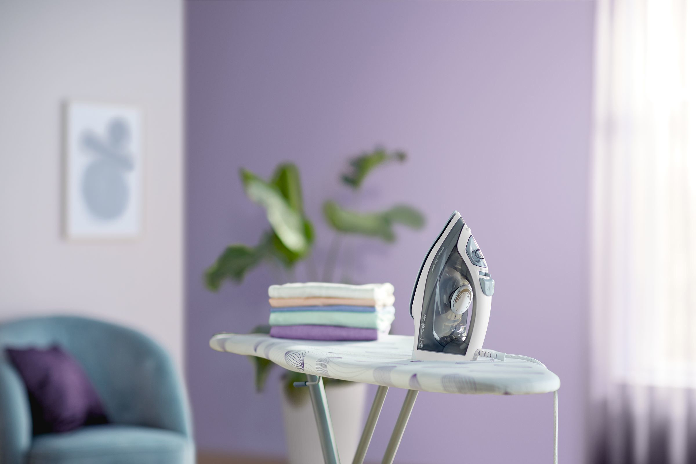 Bügelbrett mit einem Philips Bügeleisen, mit einem Stapel gefalteter Kleidung und einer lila Wand.