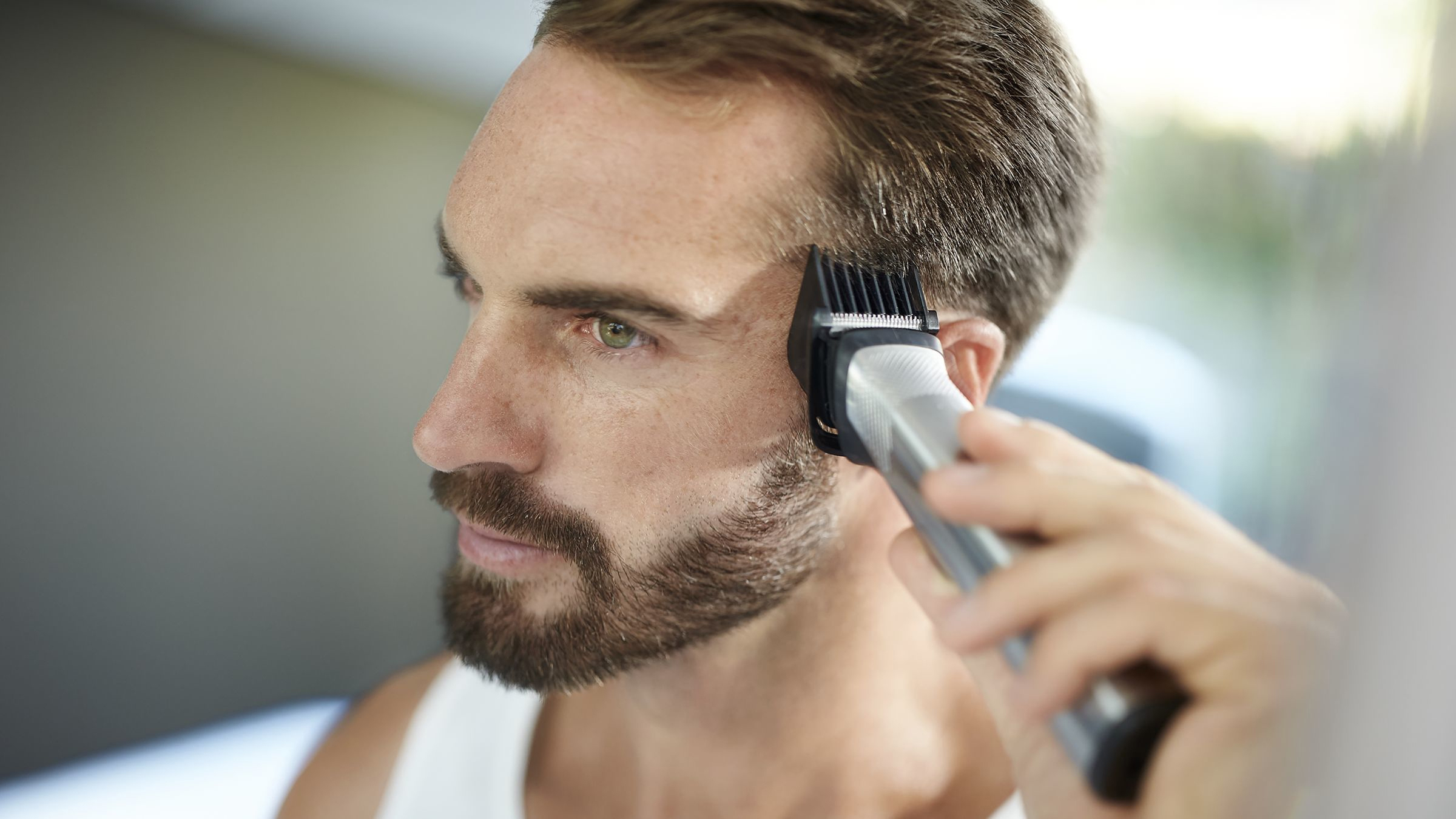 Mann rasiert seine Koteletten mit einem Trimmer. Er hat einen Bart und schaut in einen Spiegel.