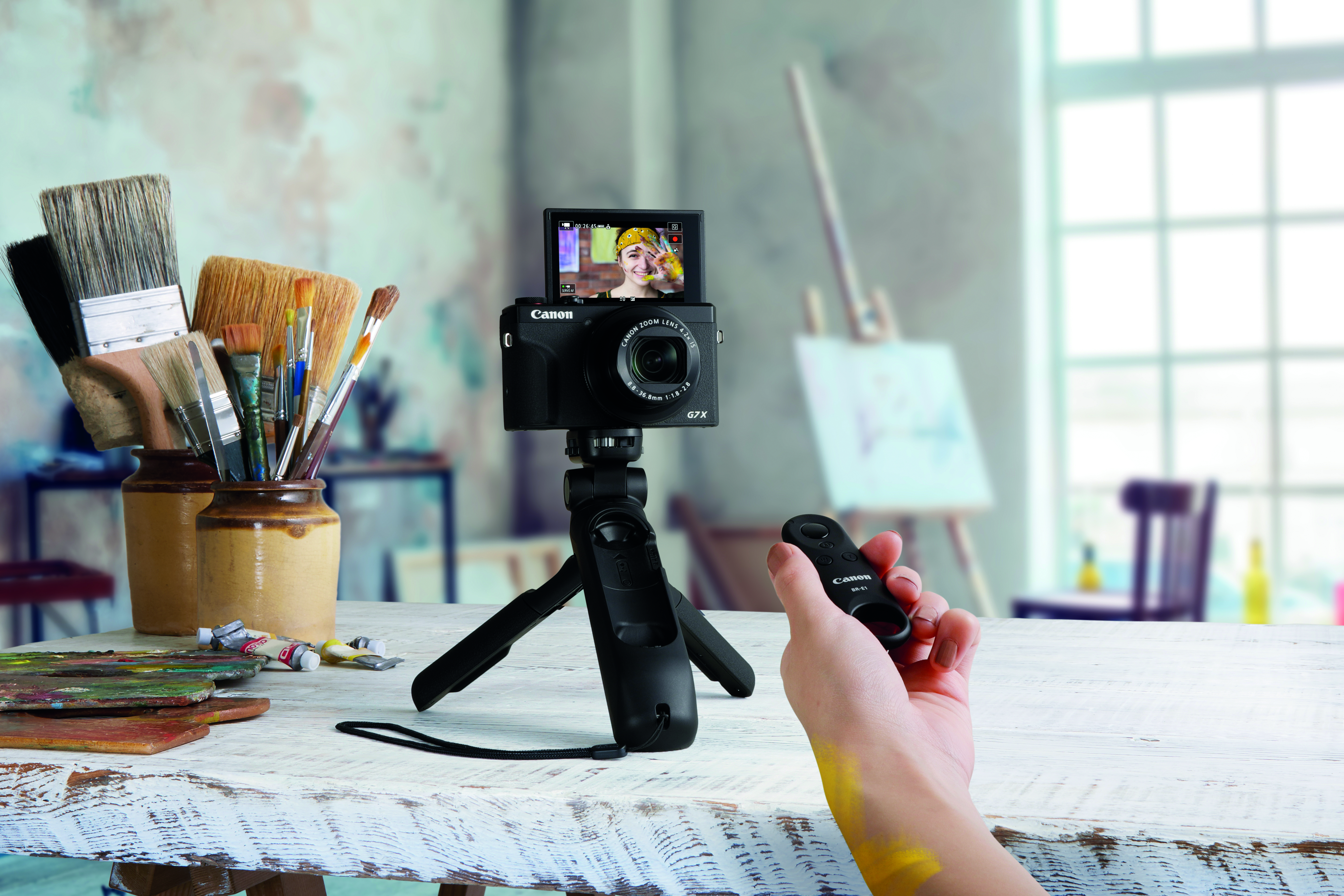 A camera on a tripod with a person holding a remote. A painting studio background.