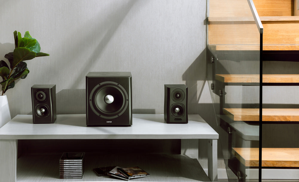 Ein Wohnzimmer-Setup mit drei schwarzen Lautsprechern auf einem weißen Tisch und einer Pflanze. Treppe sichtbar.