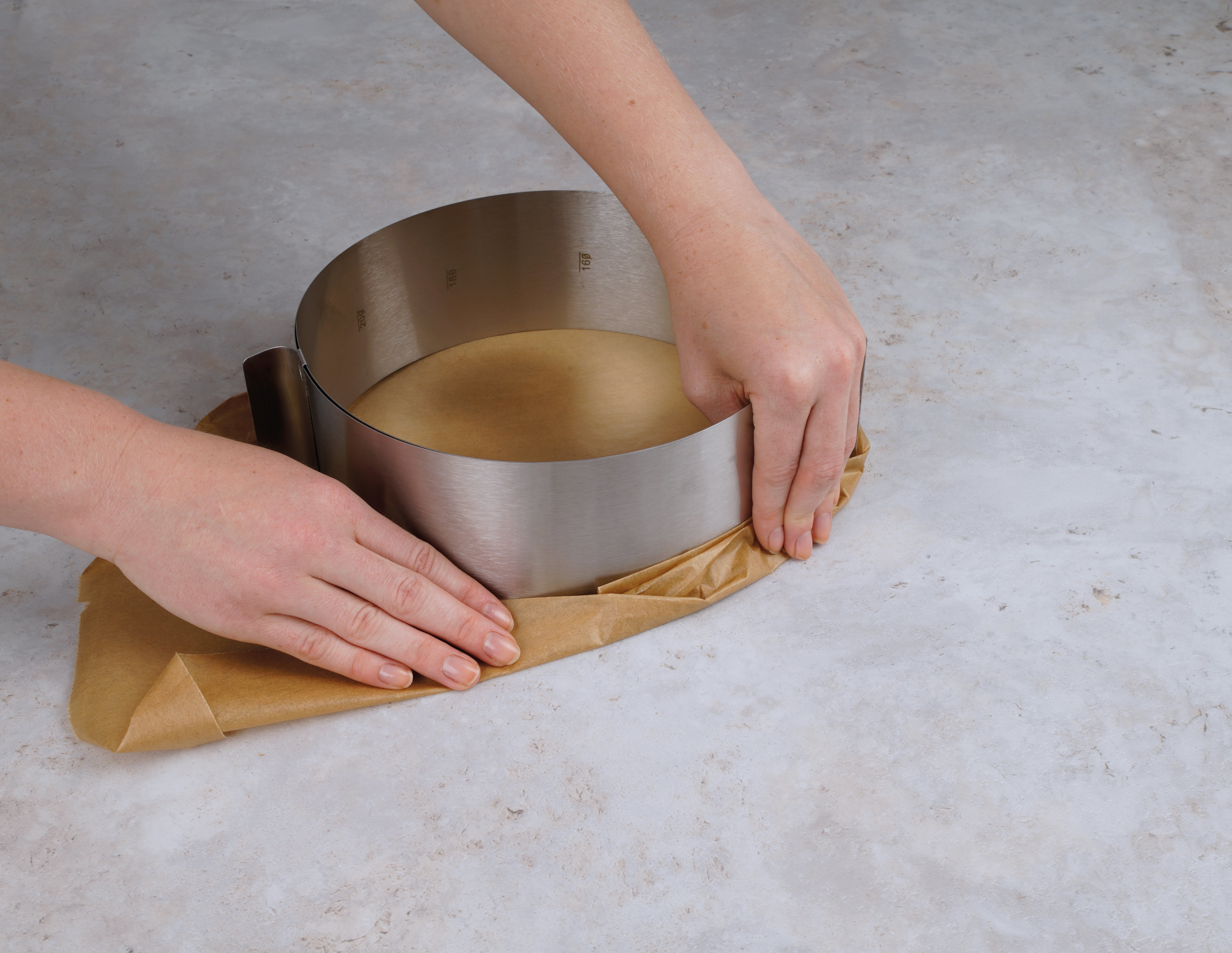 Hände einer Person, die einen Metallring um einen Kuchen auf einer hellen Oberfläche platzieren.