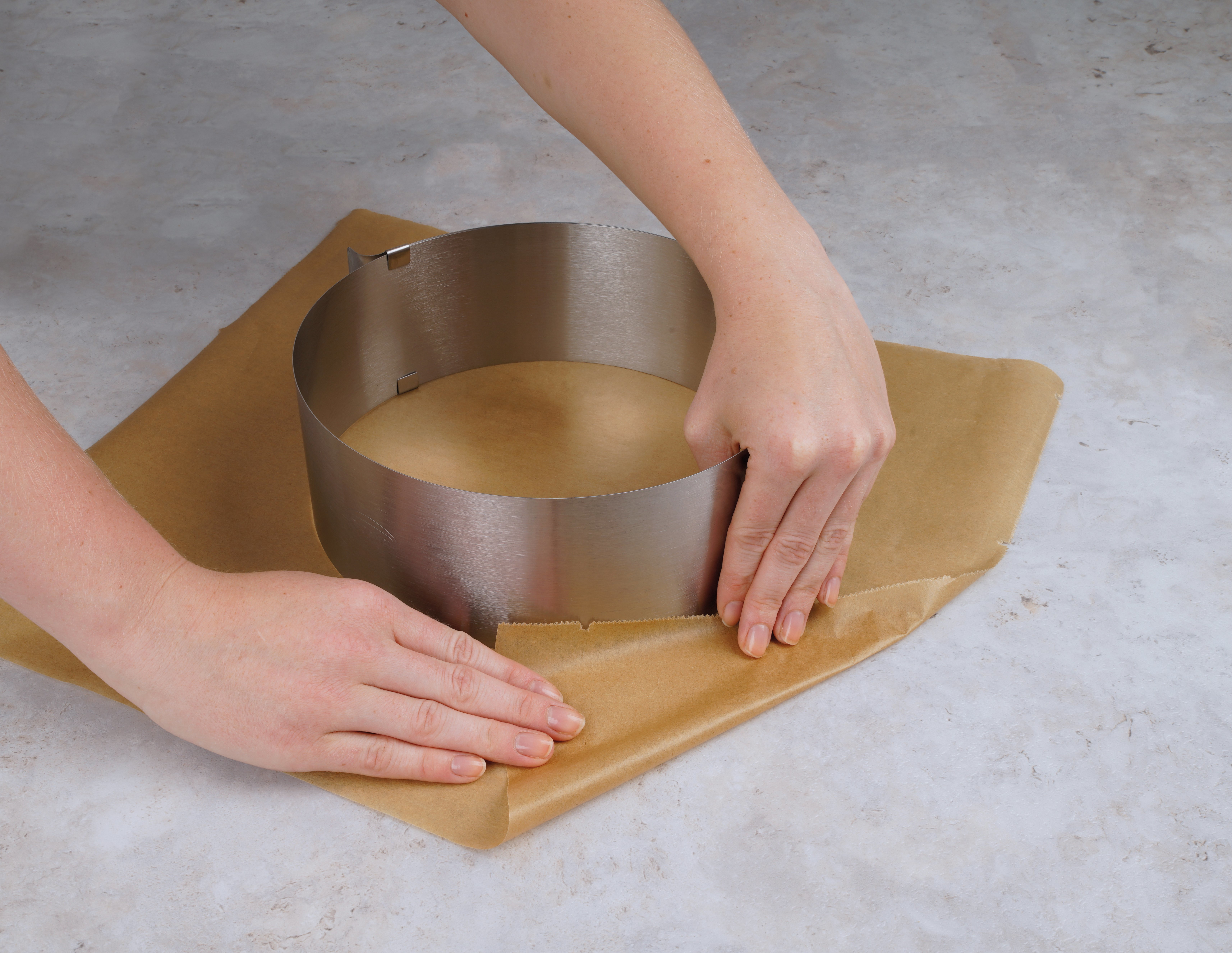 Hände legen einen Kuchenring aus Metall auf braunes Pergamentpapier auf einer hellen Arbeitsplatte.
