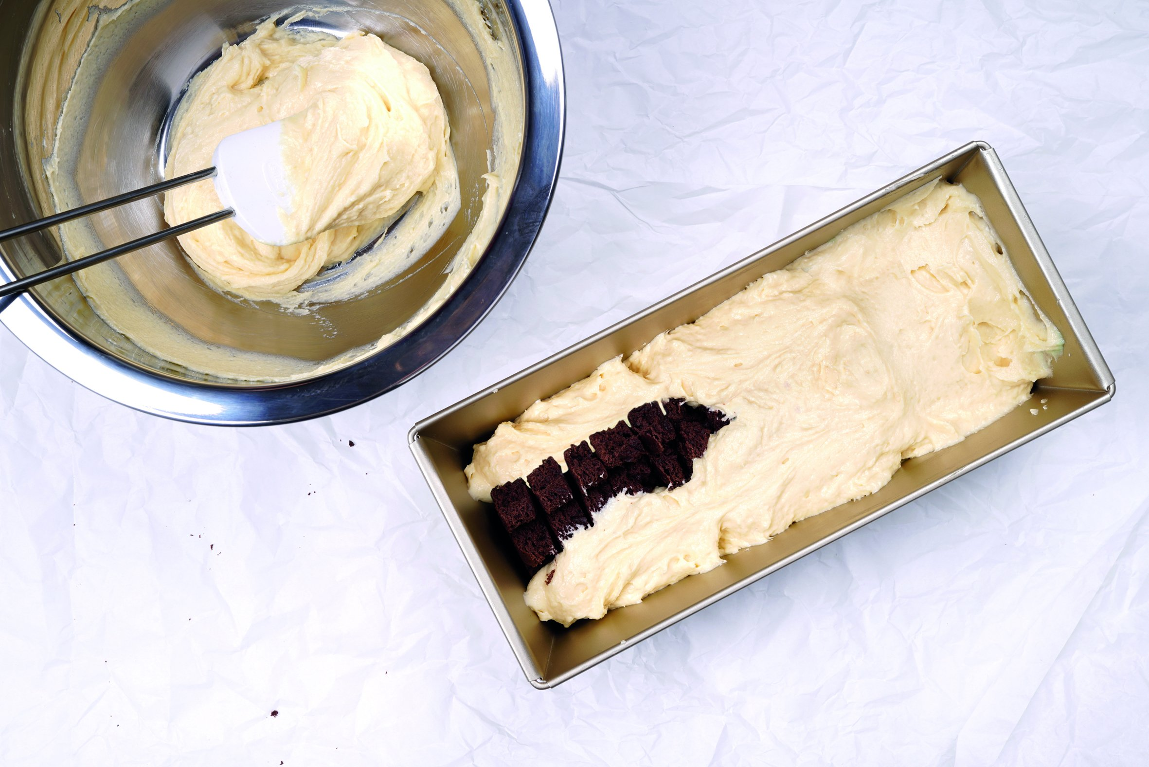 Eine Backszene zeigt eine Schüssel mit Teig, einen Spatel, eine Brotform mit Teig und Brownie-Stücken auf weißem Papier.