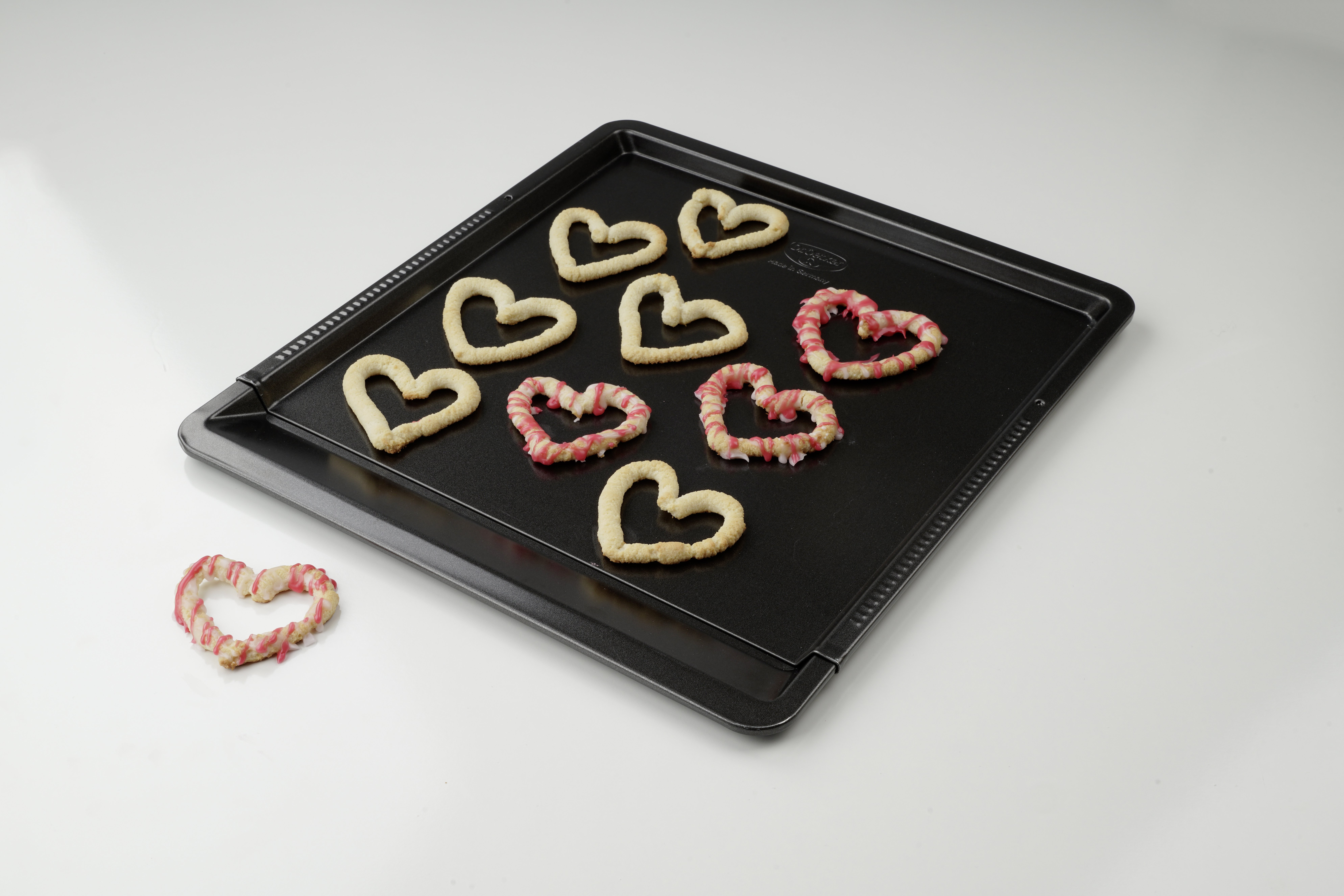 Backblech mit herzförmigen Keksen, einige mit rosa Zuckerguss verziert, freigestellt.