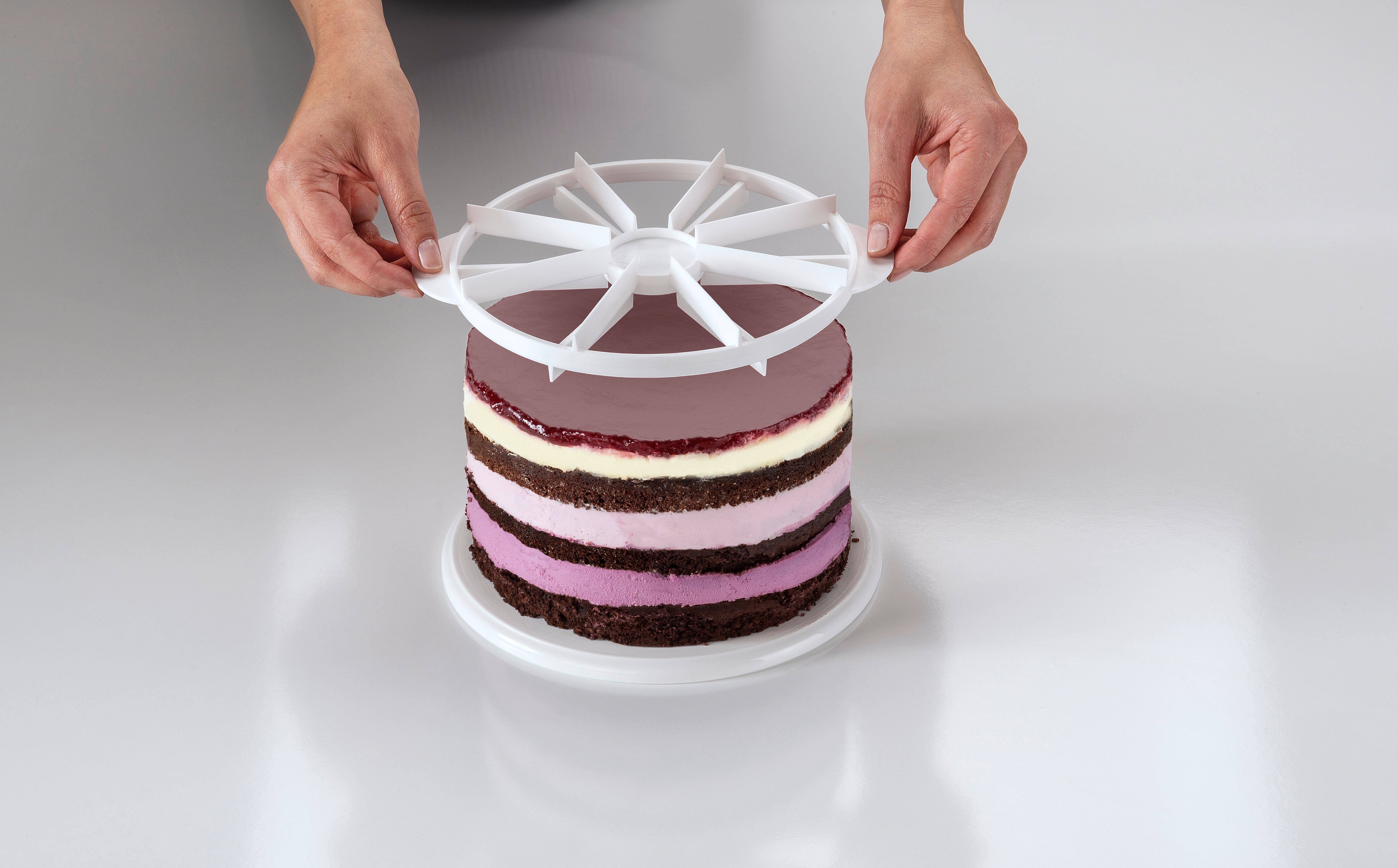Hände halten einen weißen Tortenteiler über einem geschichteten Kuchen auf einer weißen Oberfläche. Der Kuchen hat dunkle und helle Schichten.