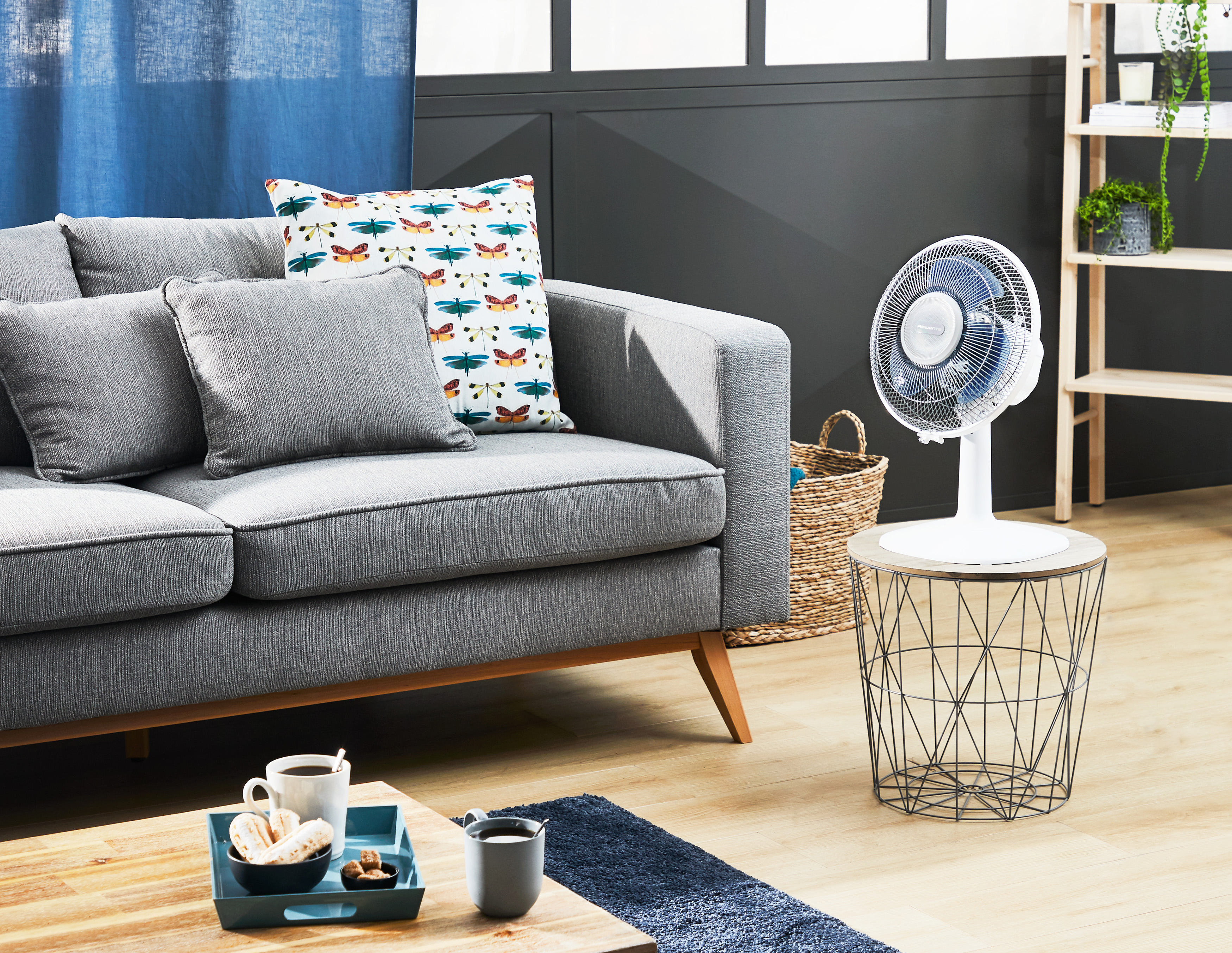 Graues Sofa mit Kissen in der Nähe eines Holztisches mit Ventilator, Kaffee und Teppichen.