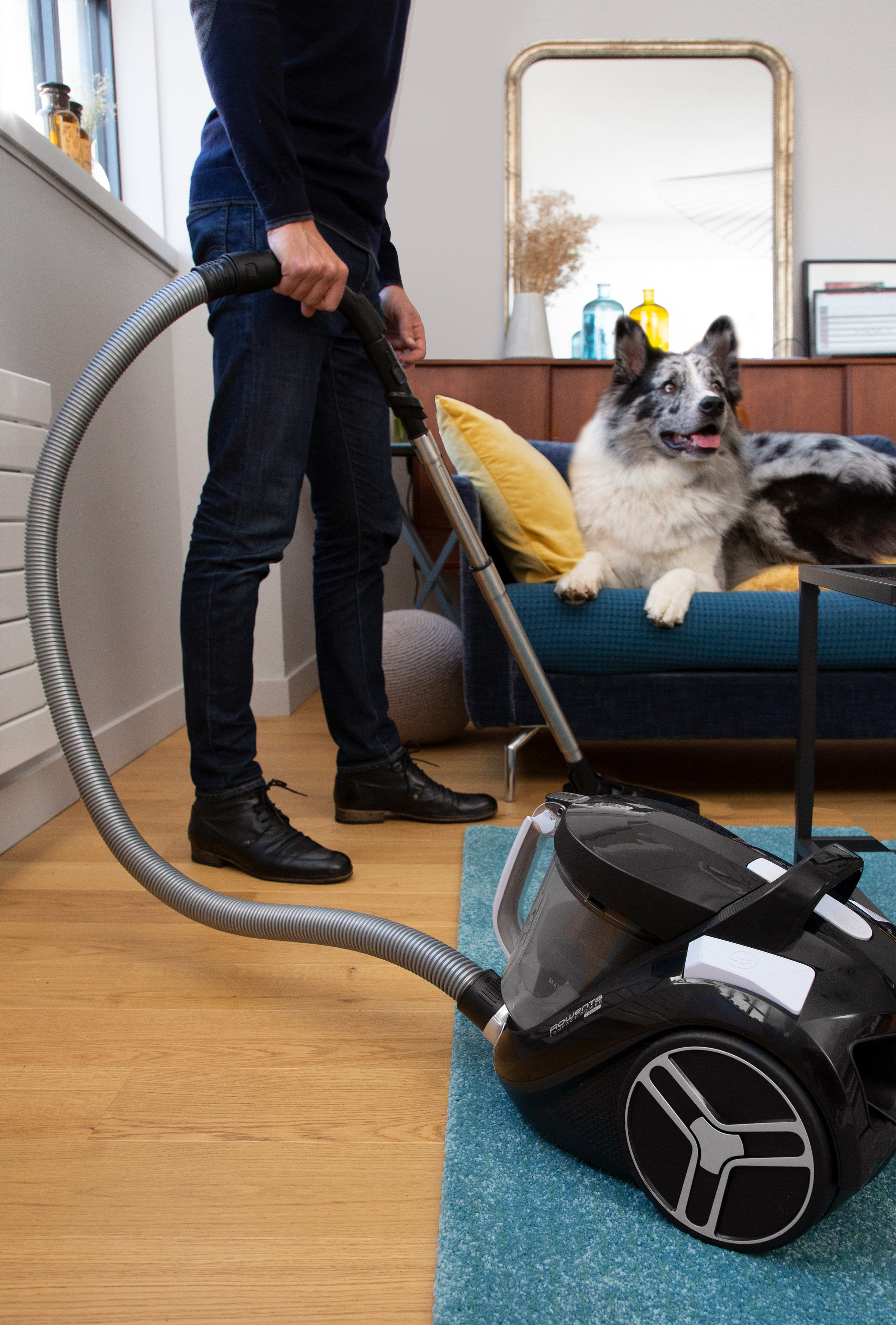 Mann saugt Wohnzimmer, Hund schaut zu.