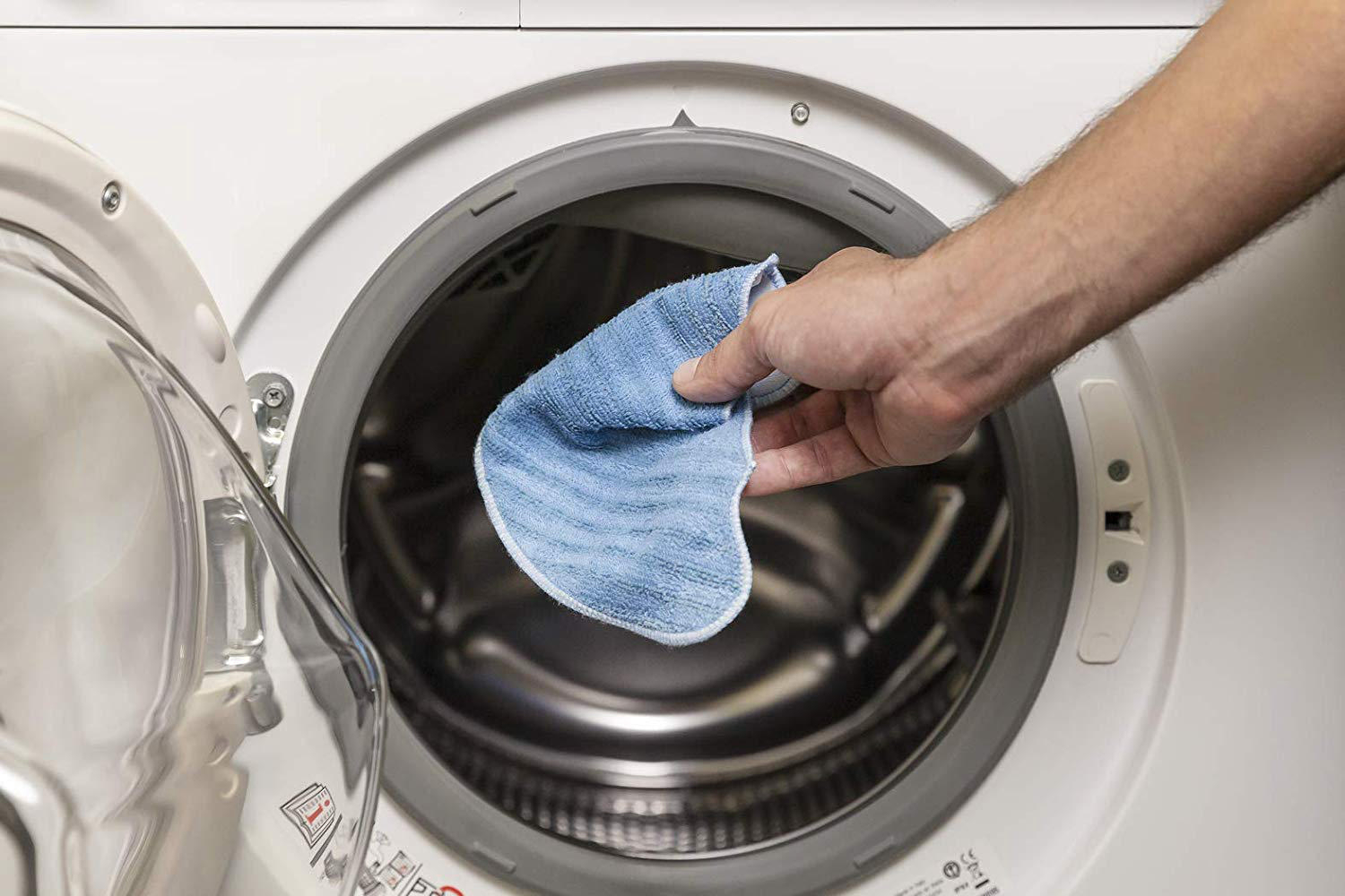 Hand, die ein blaues Tuch in die offene Tür einer weißen Waschmaschine einführt.