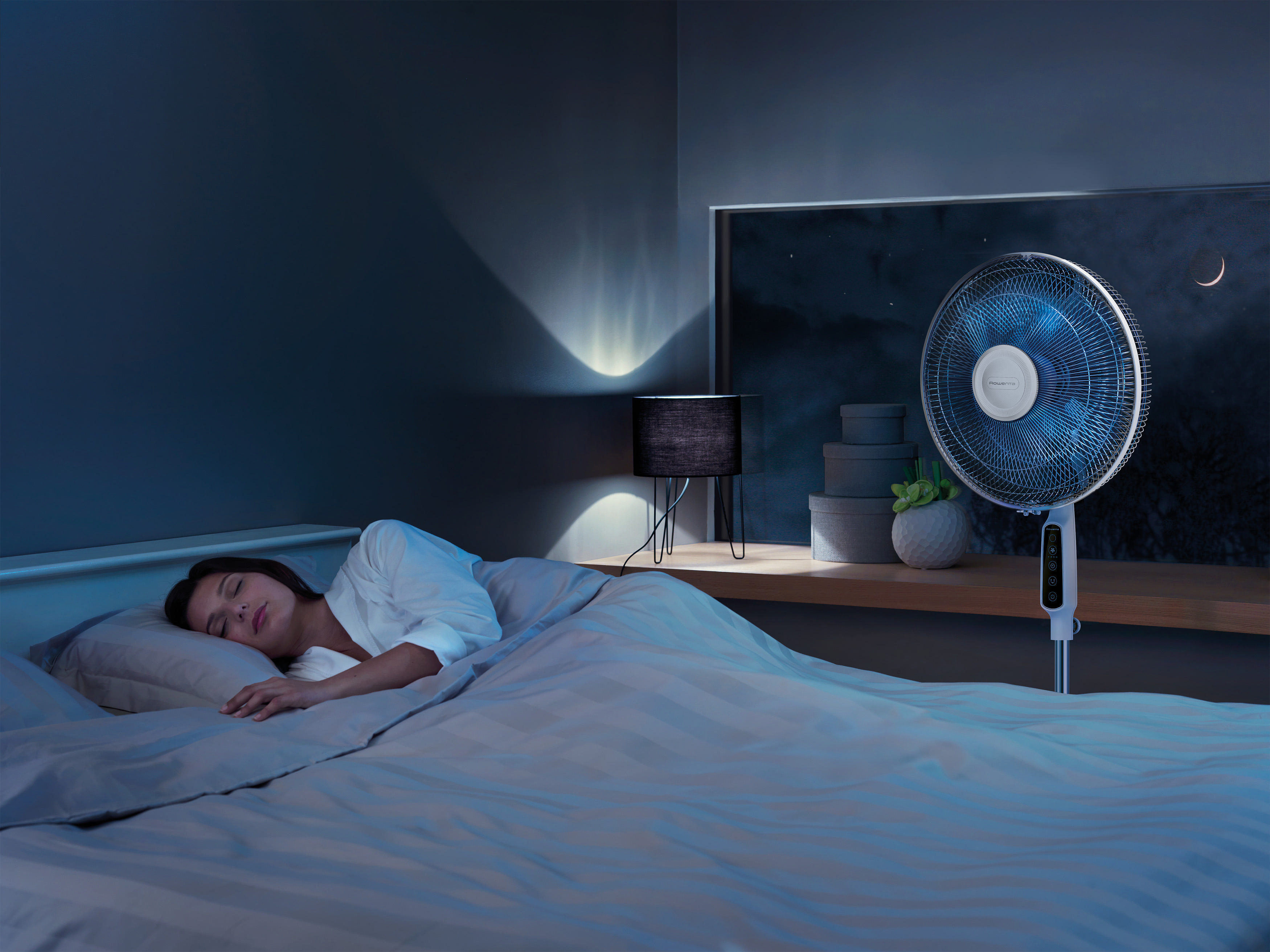 Eine Frau schläft in einem Schlafzimmer, während ein Ventilator im Hintergrund läuft, Nachtansicht.