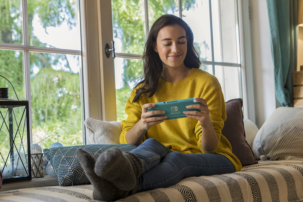 Een vrouw zit bij een raam en houdt een teal Nintendo Switch vast.