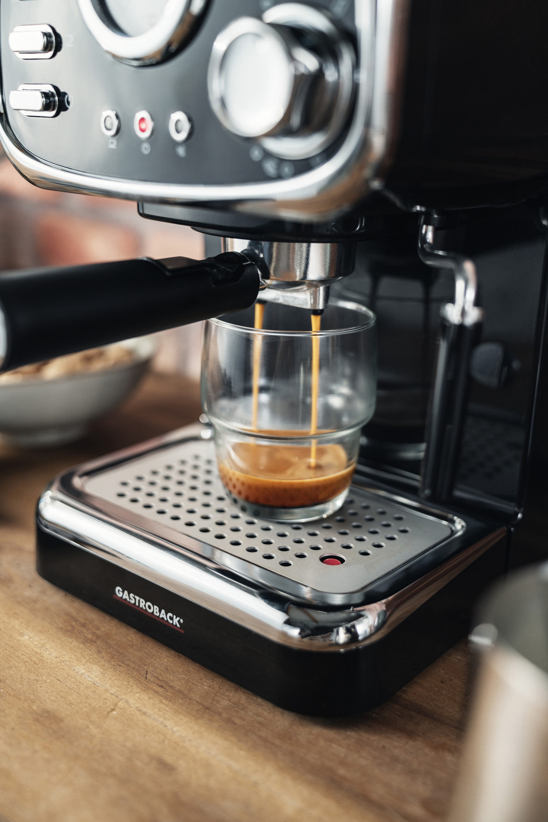 Eine Espressomaschine, die Kaffee in eine Glastasse ausgibt. Eine weiße Schüssel und Ziegelmauer im Hintergrund.