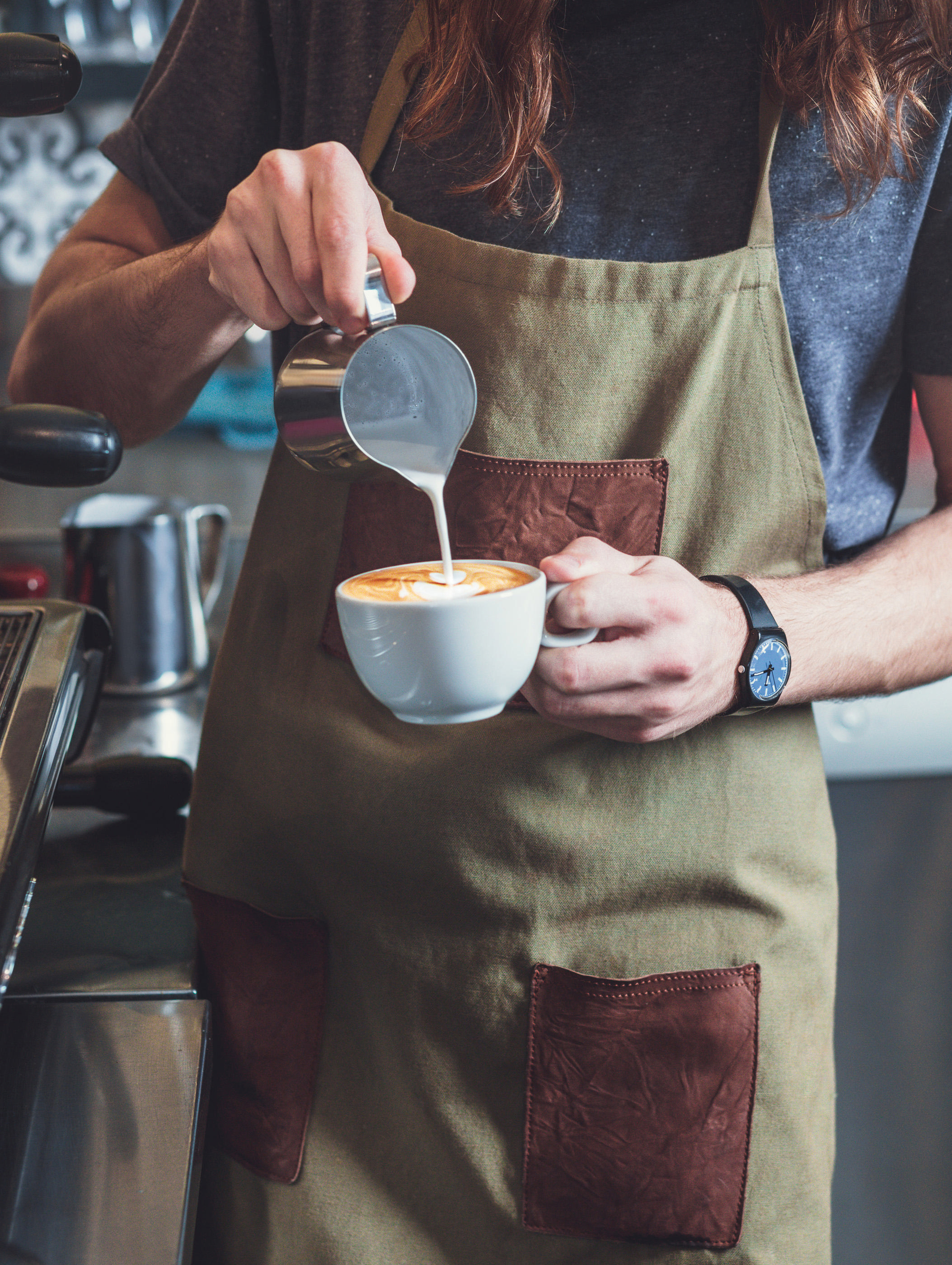 Barista gießt Milch in eine Kaffeetasse, Latte Art entsteht. Beigefarbene Schürze.