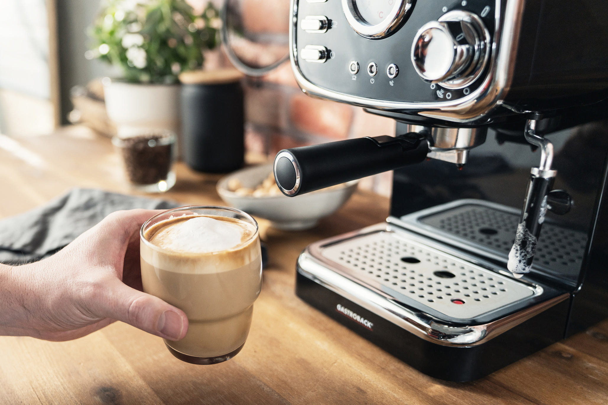 Hand hält ein Glas Latte. Kaffeemaschine rechts. Weitere Gegenstände auf Holzoberfläche.