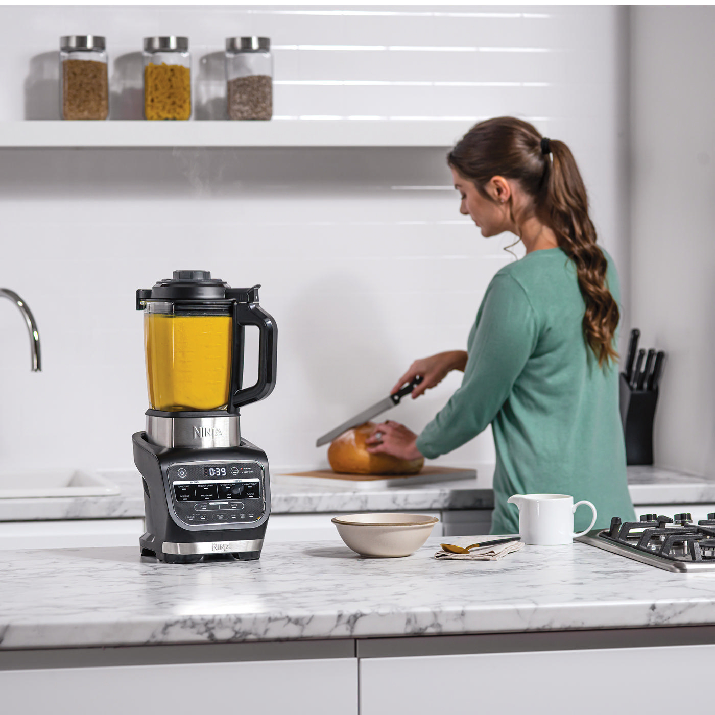 Eine Frau schneidet Brot neben einem schwarzen Mixer. Der Mixer hat eine gelbe Flüssigkeit im Inneren.