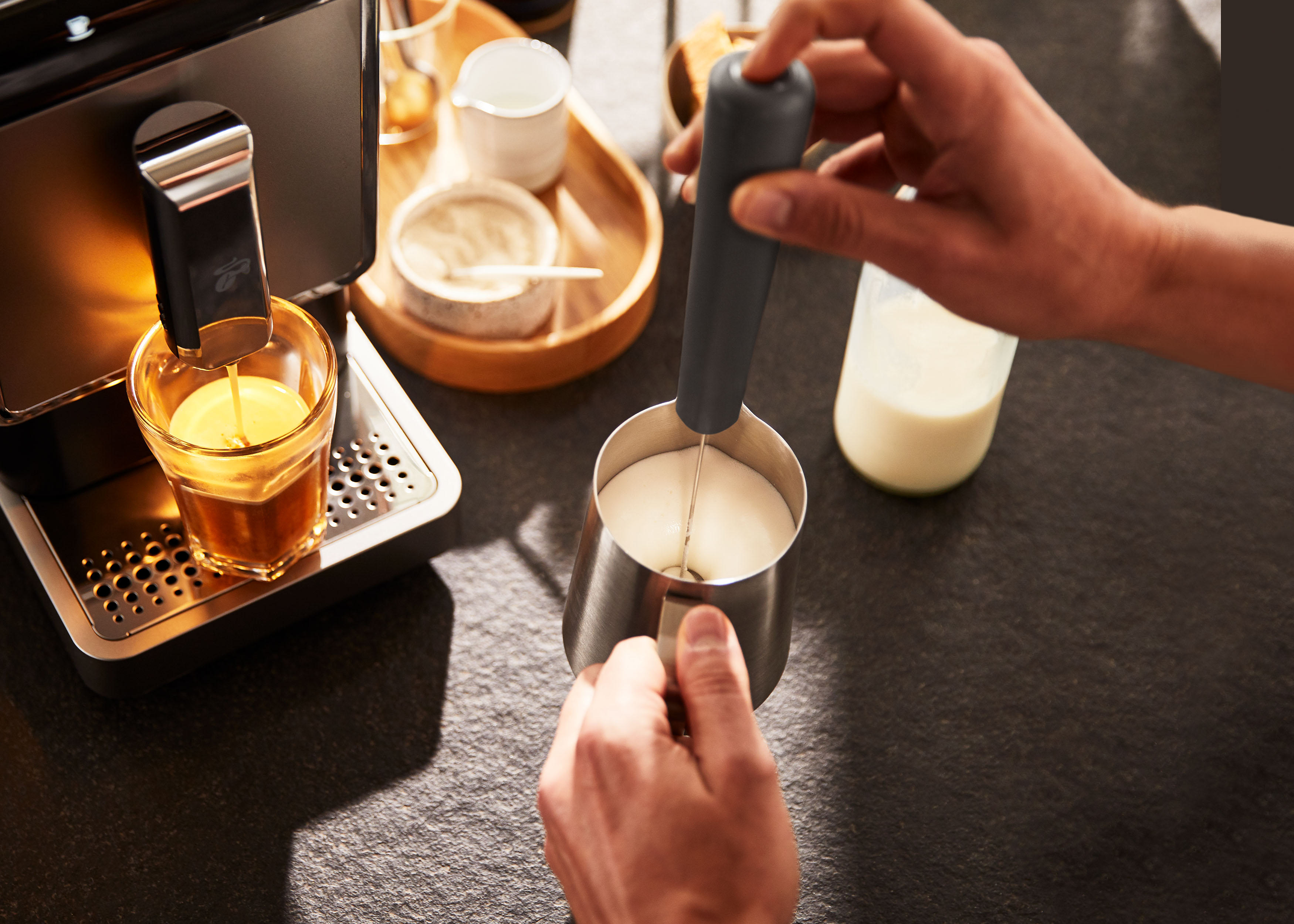 Hände schäumen Milch mit einem Aufschäumer neben einer Espressomaschine und einem Glas.