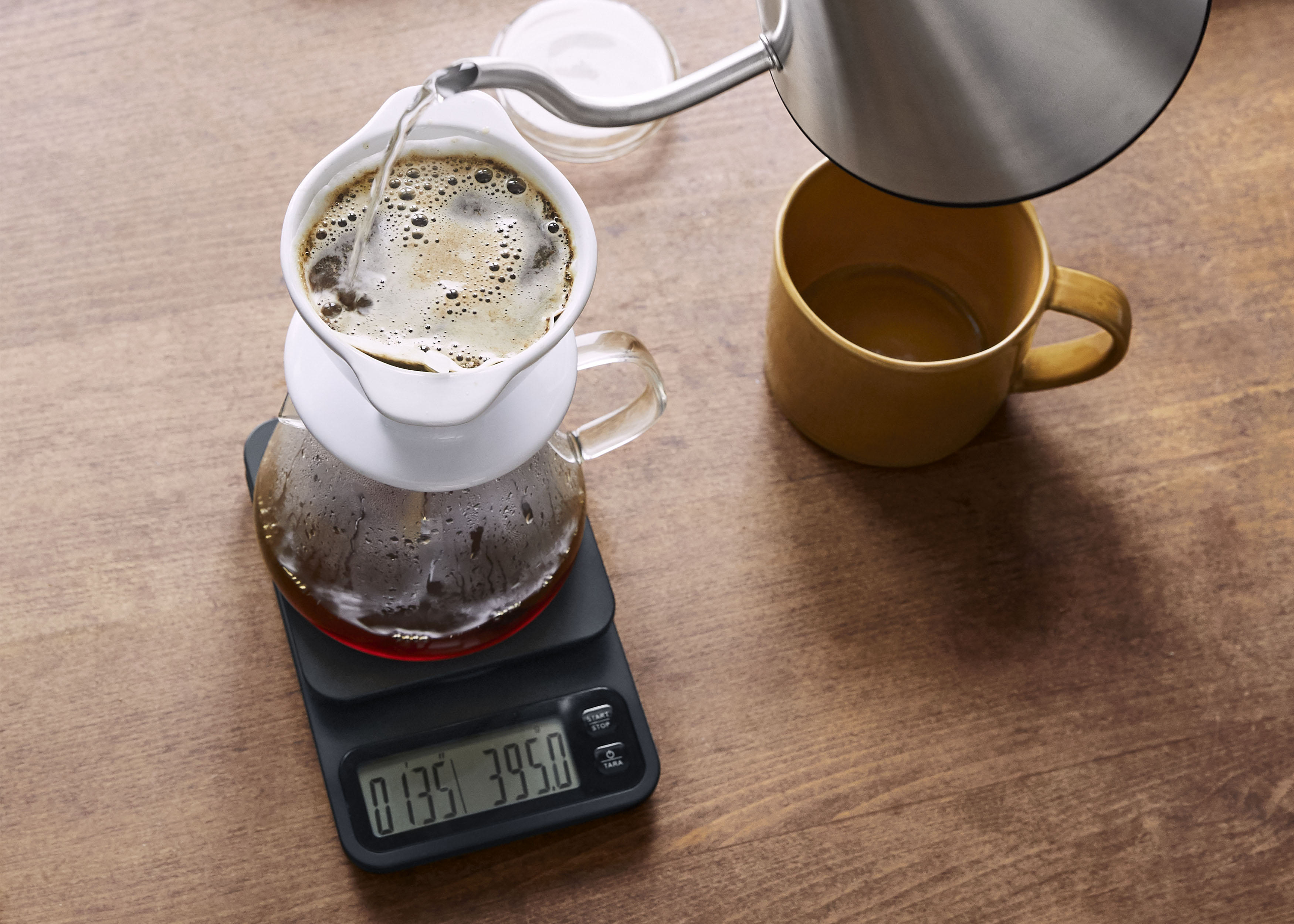 Kaffee wird gebrüht. Wasser gießt aus einem Wasserkocher in einen Pour-Over. Eine Tasse und eine Waage sind vorhanden.