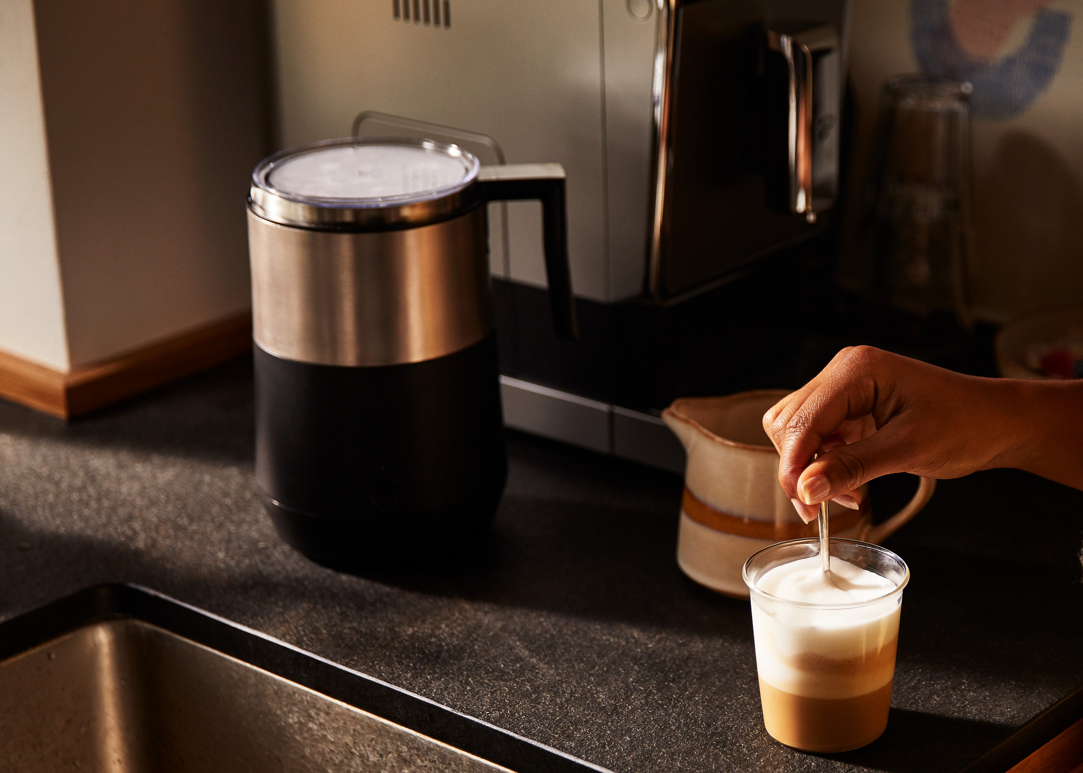 Milchaufschäumer mit Schaum, der in einem Glas verrührt wird; Kaffeemaschine im Hintergrund.