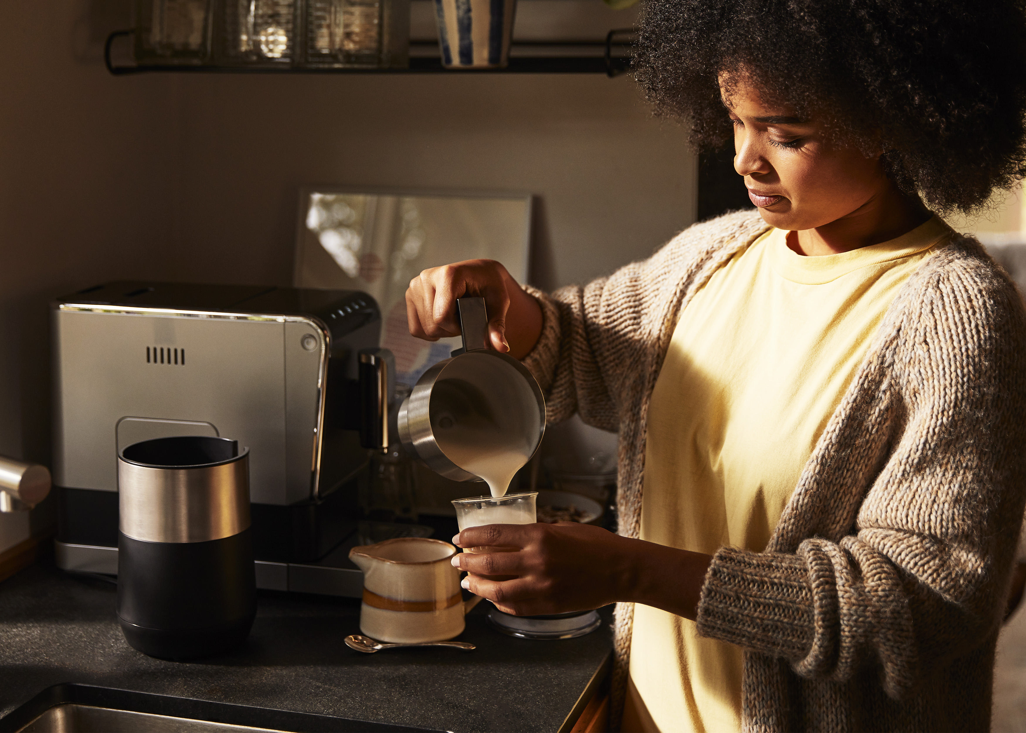 Frau gießt Milch in eine Tasse, in der Nähe von Kaffeemaschine und Becher, in der Küche.