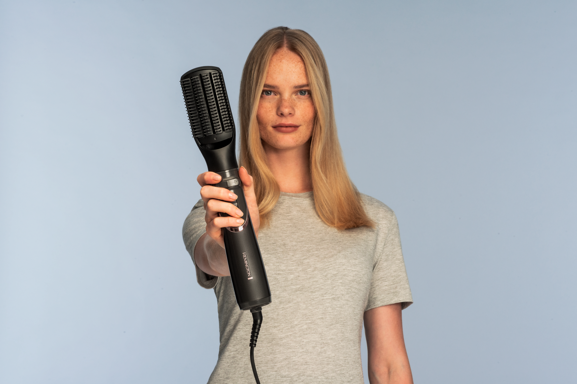 Eine Frau mit blonden Haaren hält einen schwarzen Haarstyler in Richtung Betrachter.