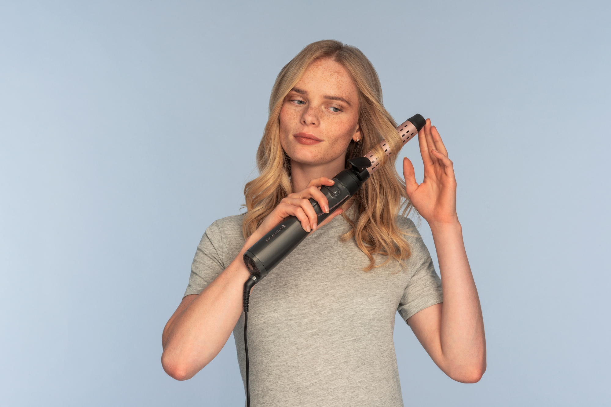 Eine Frau mit blondem Haar benutzt ein dunkelgraues Remington-Haarstyling-Tool. Sie trägt ein graues T-Shirt, hellblauer Hintergrund.