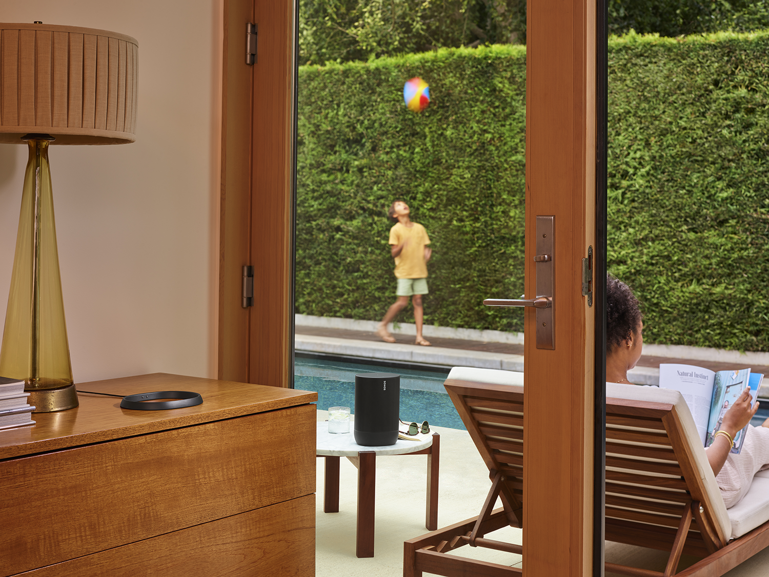A person reads a book on a lounge chair near a speaker by a pool, another plays ball.