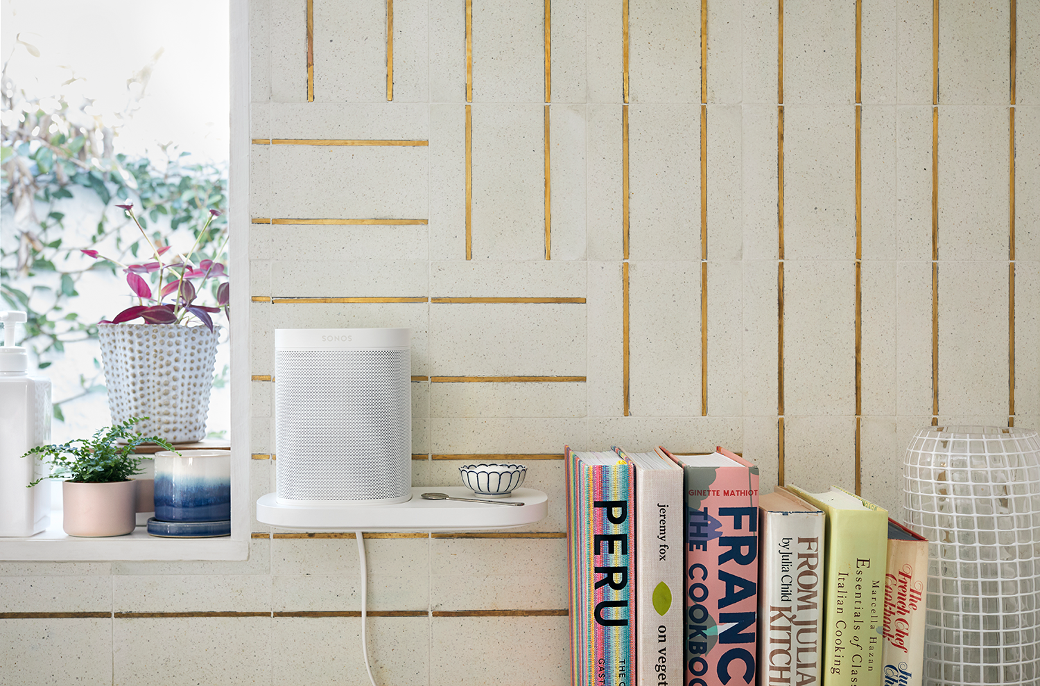 A white Sonos speaker on a shelf with books and decorations, near a window and tile wall.
