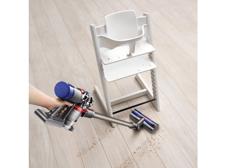 Eine Hand benutzt einen Dyson-Staubsauger auf einem Holzboden. Ein weißer Hochstuhl ist im Hintergrund. Schmutz auf dem Boden.