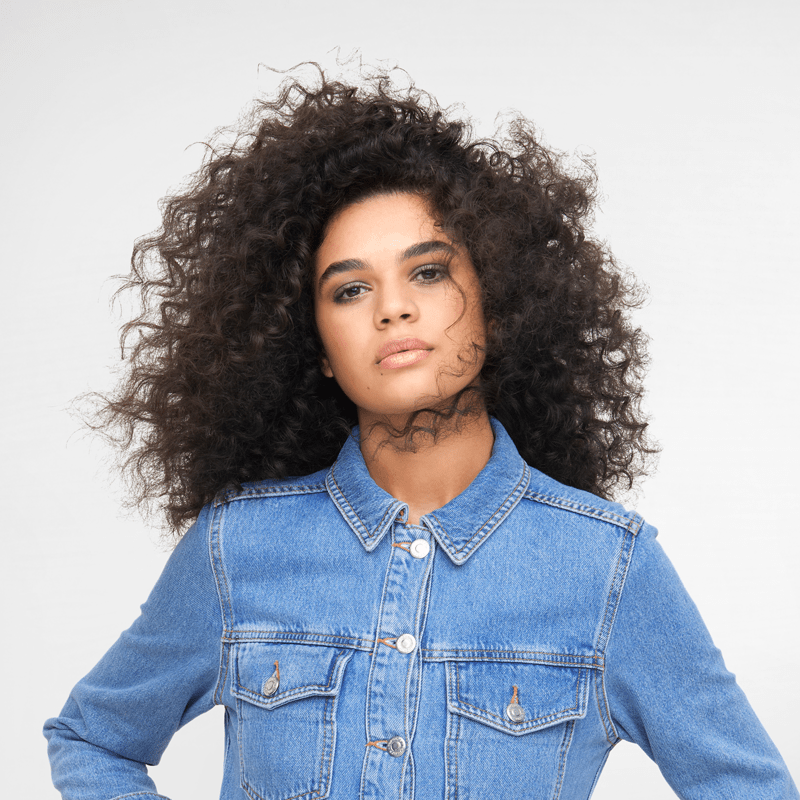 Frau mit lockigem Haar, Denim-Jacke, vor weißem Hintergrund, Blick nach vorne.