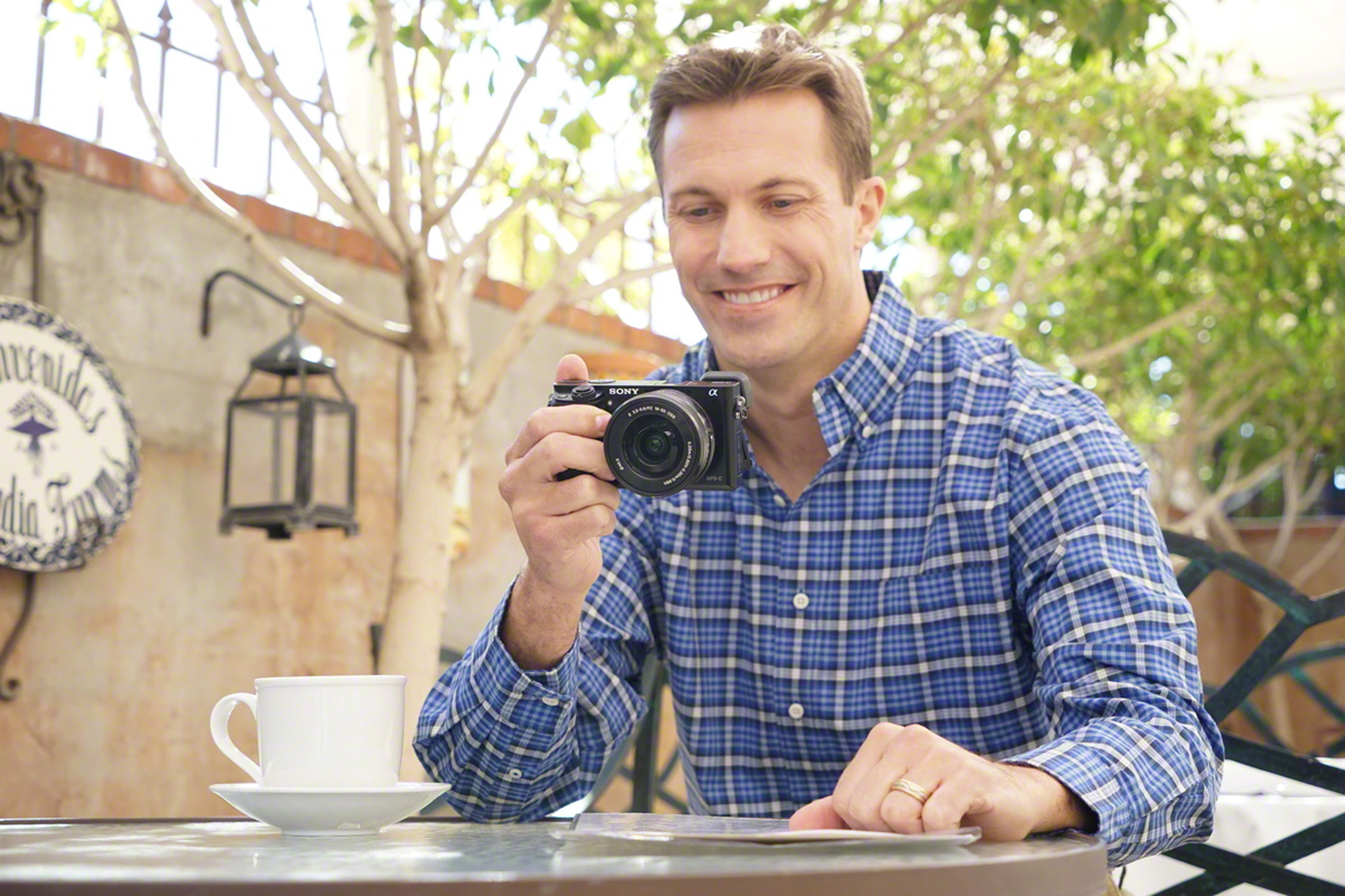 Mann sitzt am Tisch, hält Kamera, lächelt.