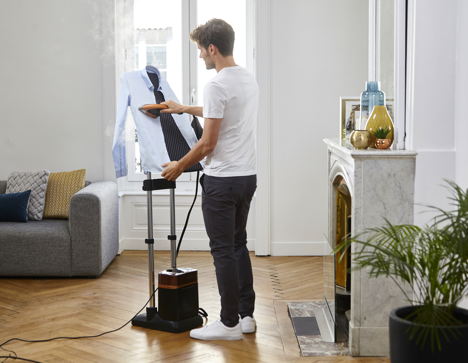 Homme repassant une chemise bleue sur une machine à repasser à vapeur dans un salon. Une plante et un miroir.