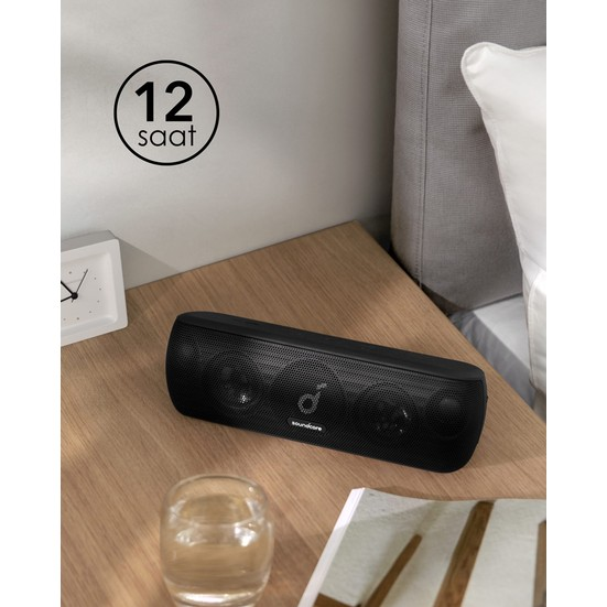 Ahşap bir yüzeyde siyah bir Soundcore hoparlör. Yakınında bir saat ve bir bardak su var.