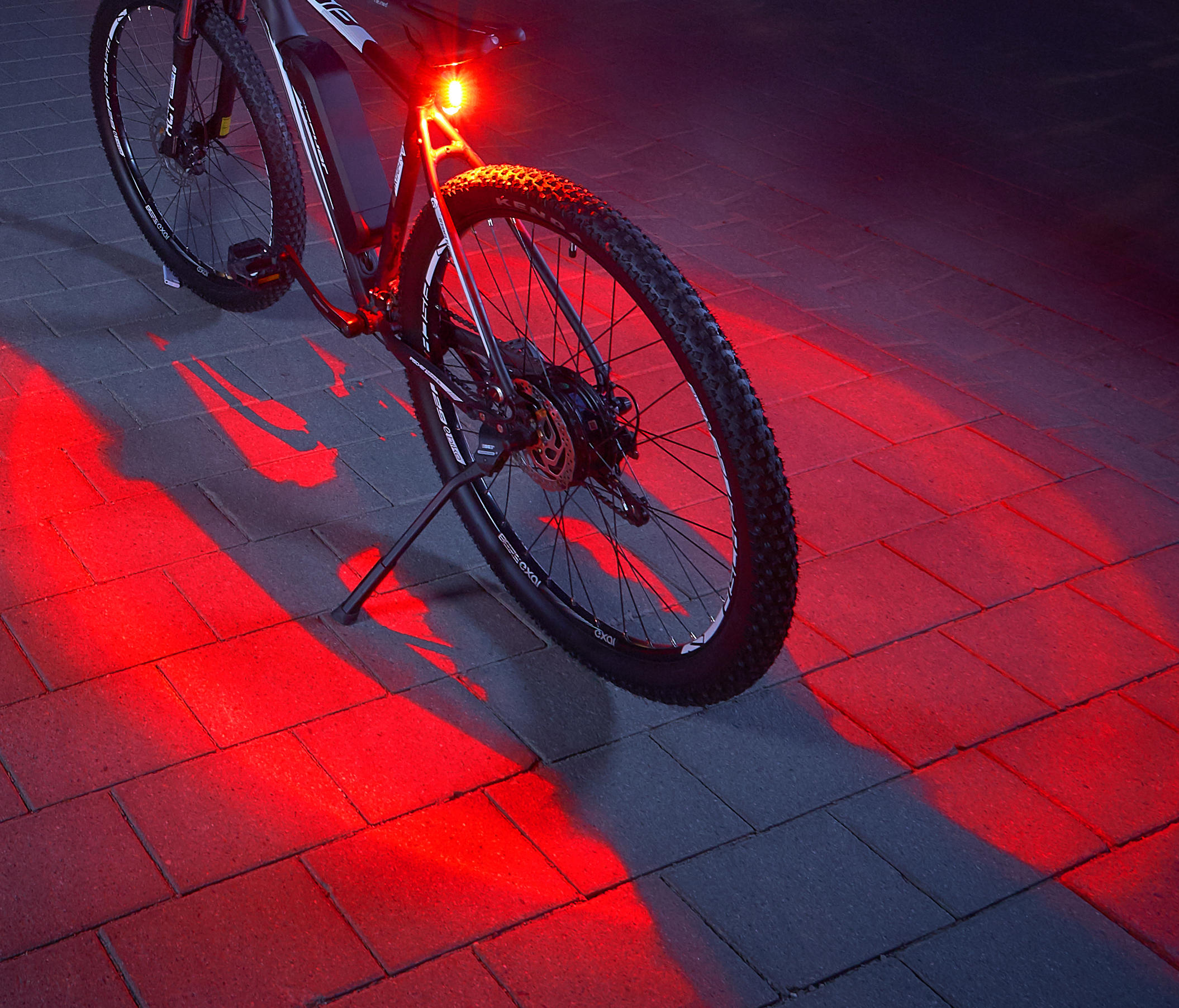 Schwarzes Fahrrad mit rotem Licht am Hinterreifen. Das Licht reflektiert sich auf dem Bürgersteig.