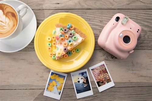 Una cámara Instax rosa, una porción de pizza con cereales y tres fotos sobre una mesa de madera.