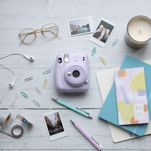 Una cámara Instax morada con accesorios sobre una superficie de madera blanca. Se ven bolígrafos, gafas y fotos.