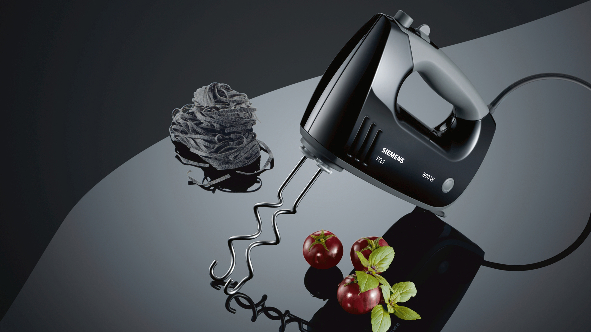 Schwarzer Handmixer mit Pasta, Tomaten und Basilikum auf einer reflektierenden Oberfläche.