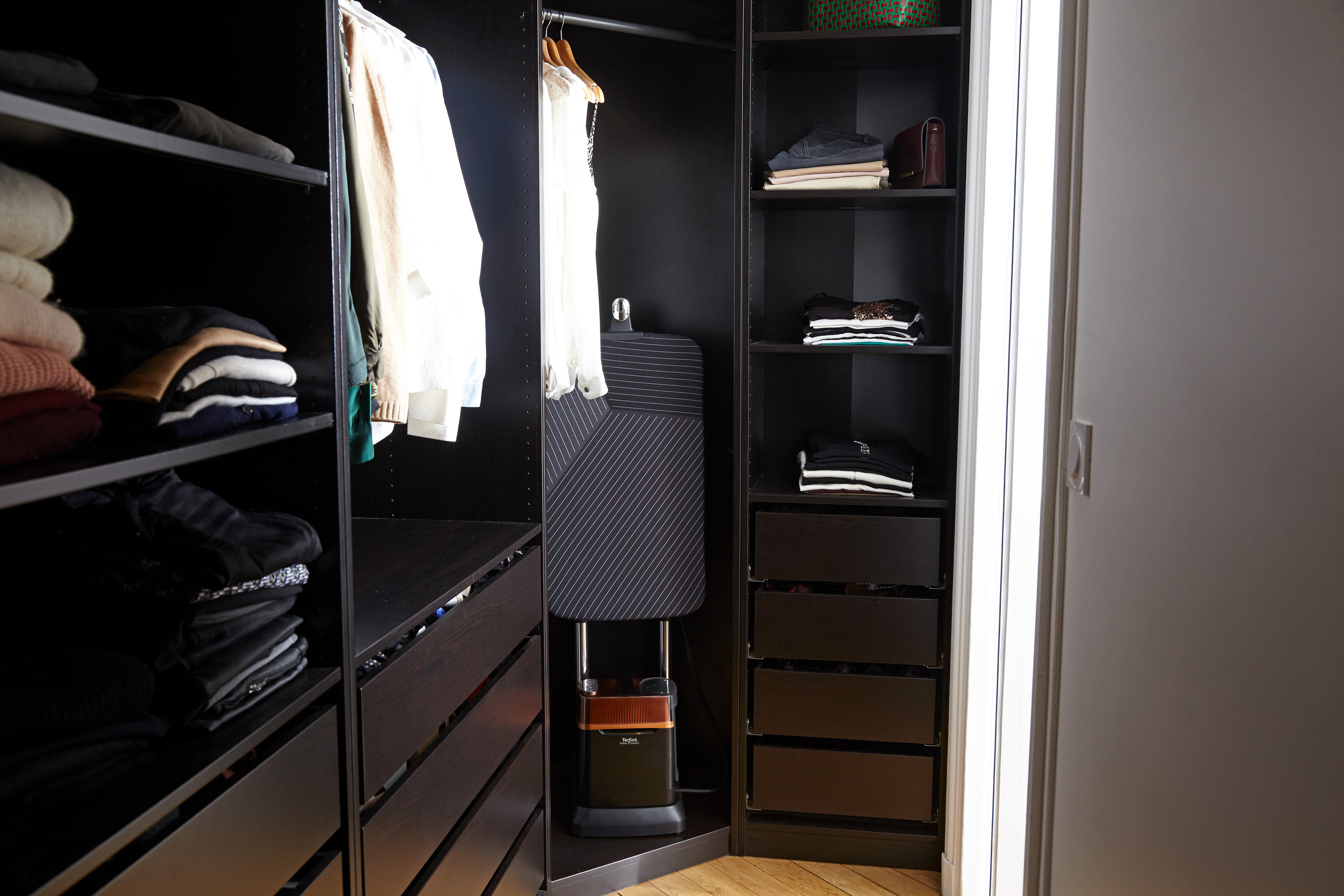 Schwarzer Schrank mit Kleidung, Regalen, Schubladen und einem Dampfgarer. Weißes Hemd hängt. Licht von rechts.