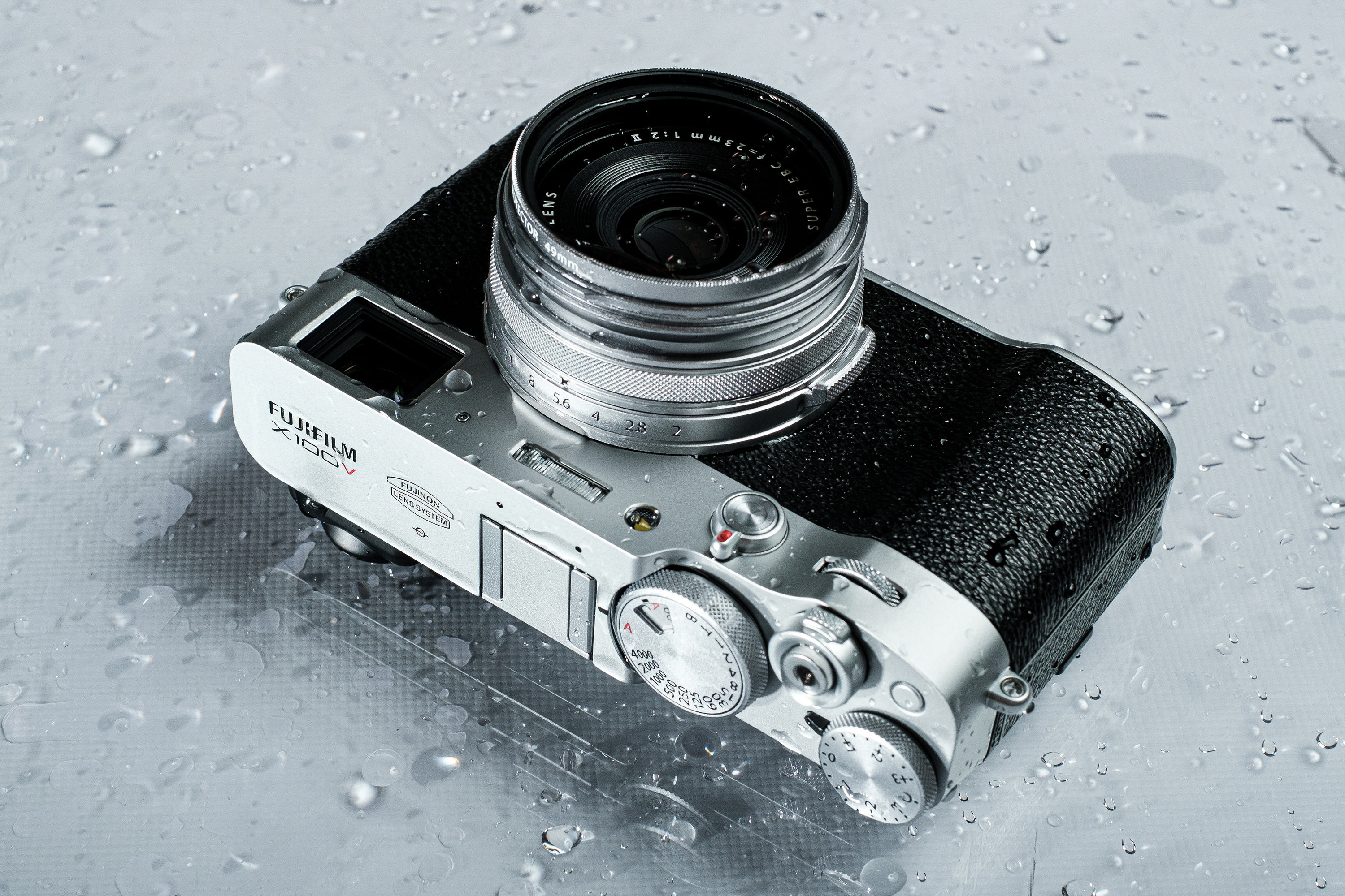 Draufsicht auf eine Fujifilm X100V Kamera mit Objektiv auf einer nassen, glänzenden Oberfläche.